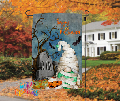 A Halloween-themed garden decoration featuring a gnome wrapped in bandages, a carved jack-o-lantern, and a gravestone with the word "RIP" on it, set against a backdrop of autumn foliage.