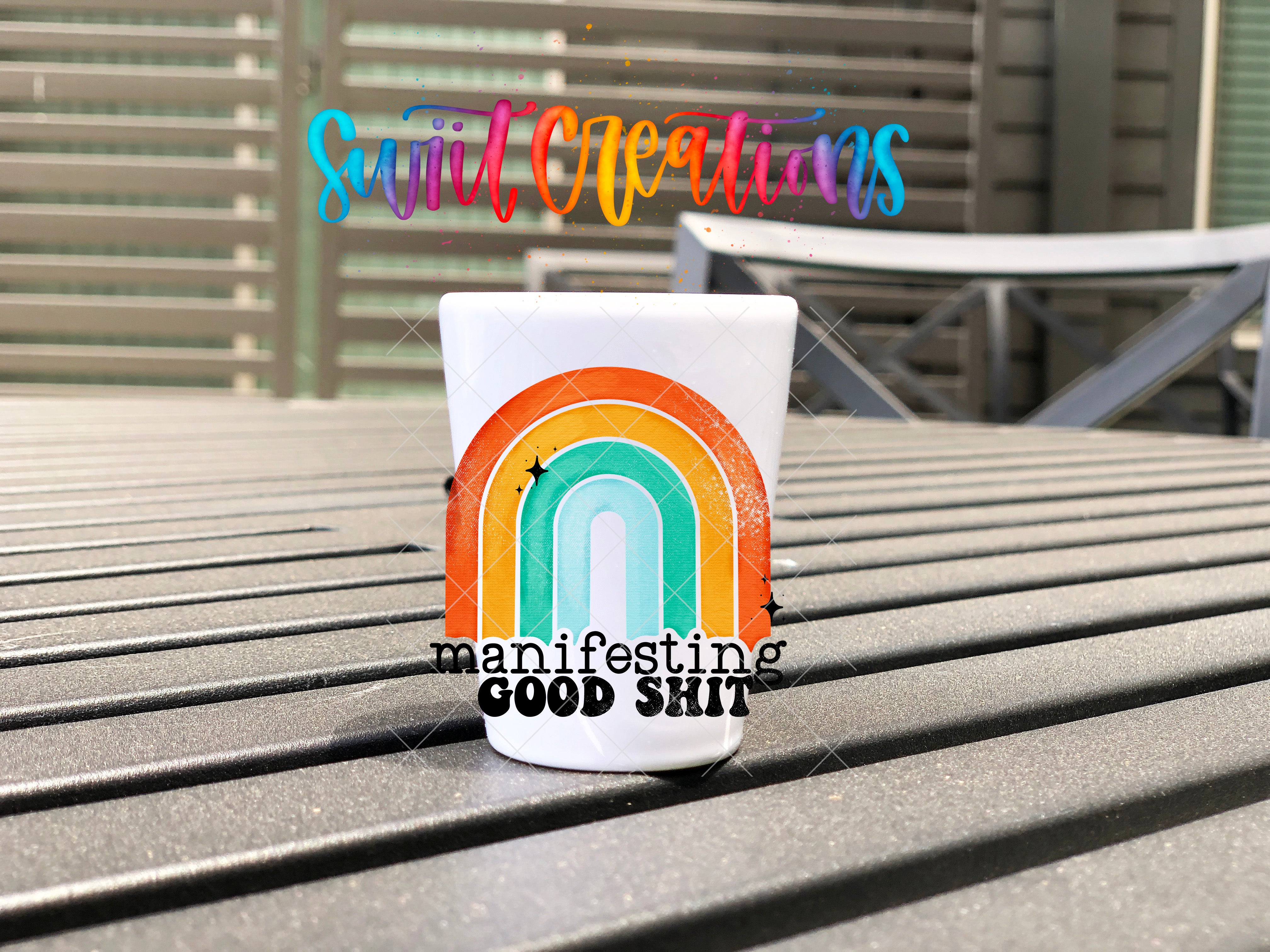 A white mug with a colorful rainbow graphic and the text "manifesting GOOD SHIT" is placed on a metal bench.