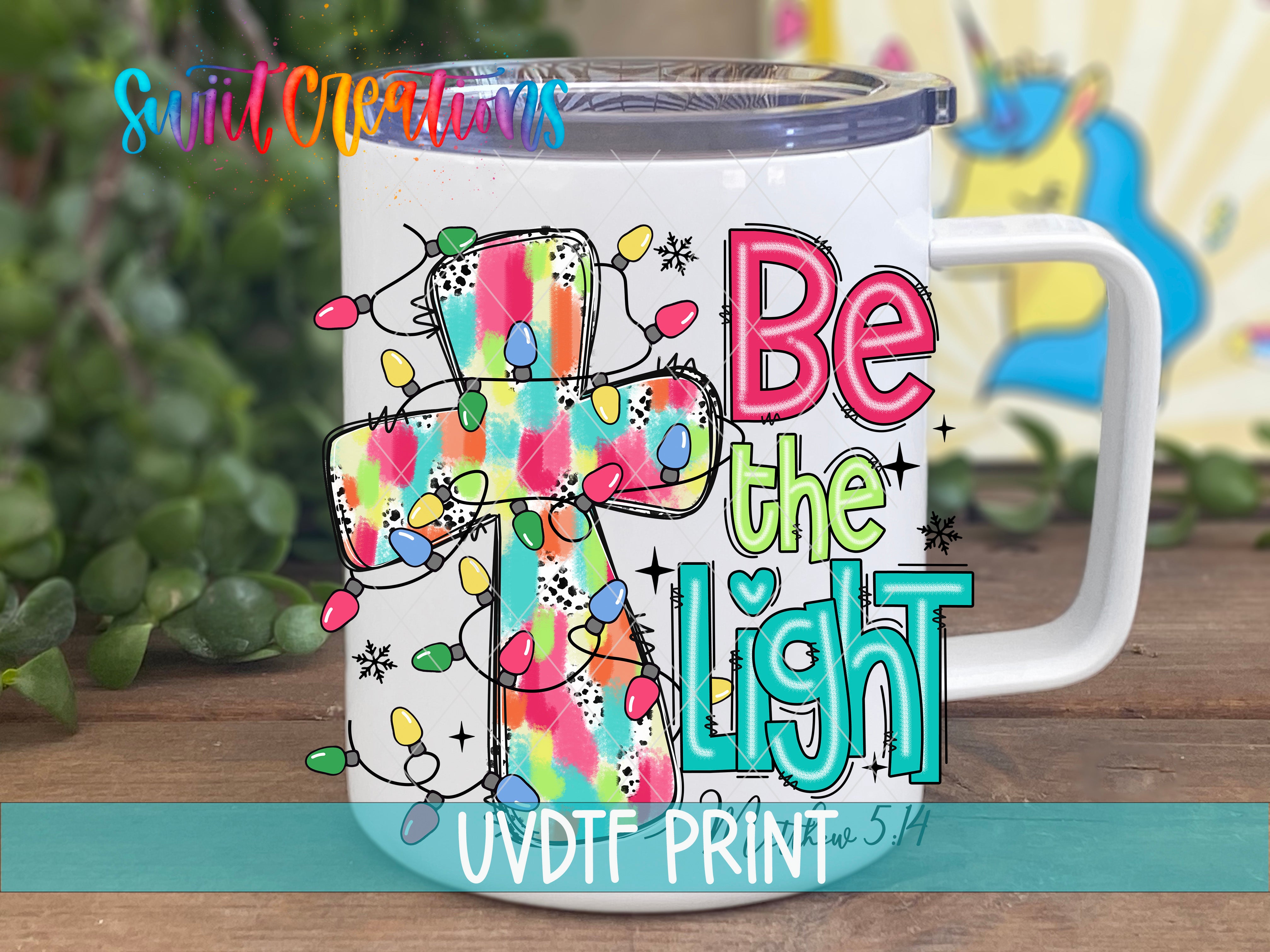 A white mug with a colorful, abstract design featuring a cross and the text "Be the Light" printed on it.