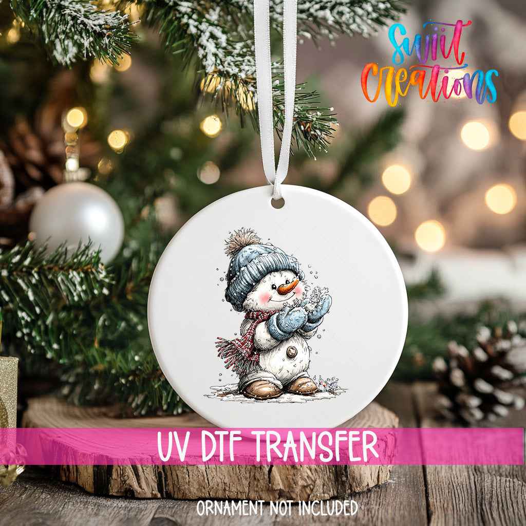 A white ornament with a snowman design hangs on a Christmas tree, surrounded by pine branches and twinkling lights.