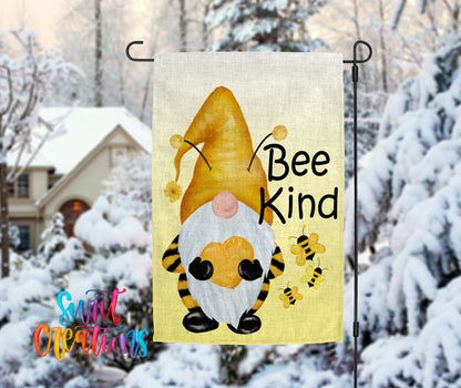 A garden gnome with a yellow hat and a bee on its back, along with the text "Bee Kind" is displayed on a flag, set against a snowy background with a house and trees in the distance.