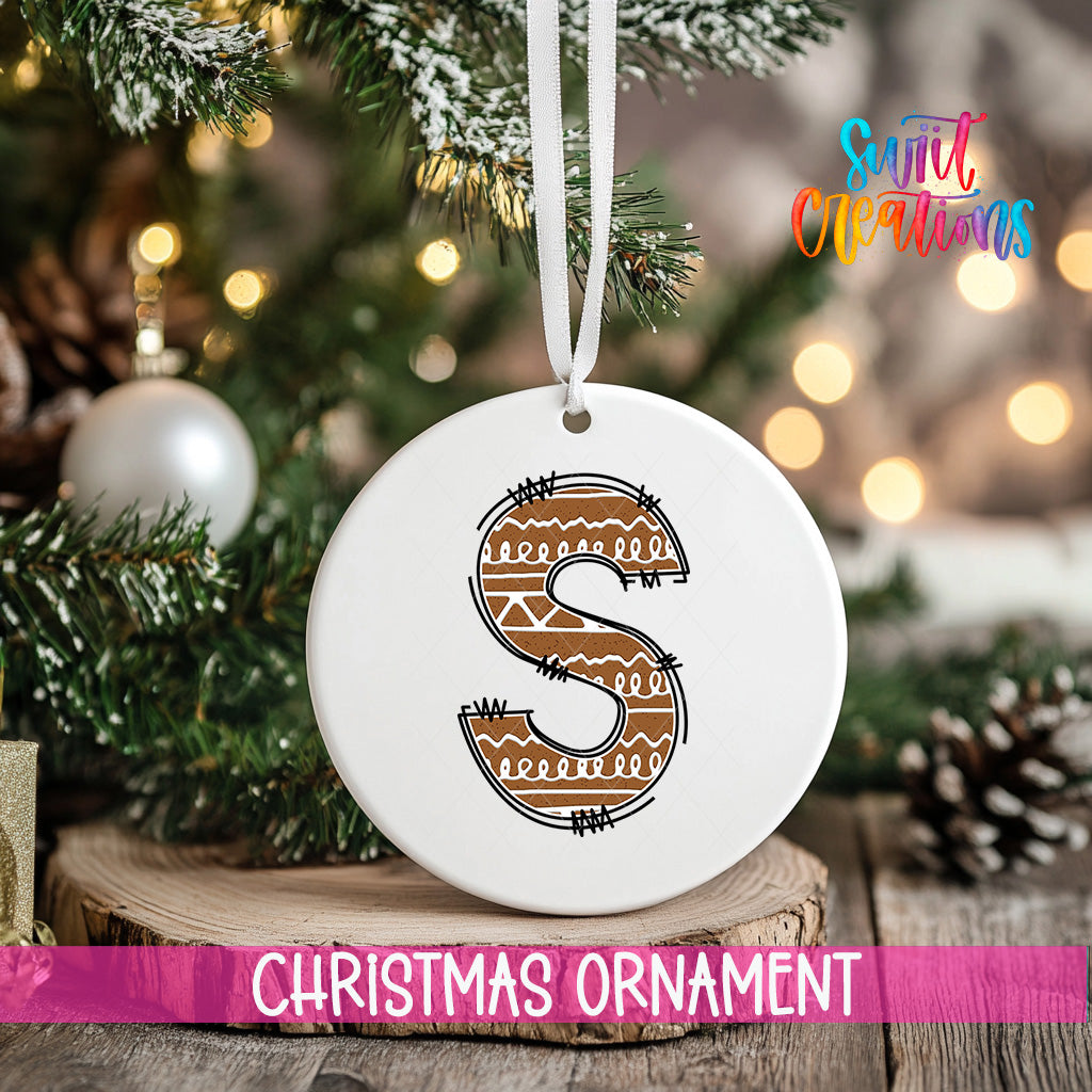 A white Christmas ornament with a brown letter "S" design hangs from a tree branch, surrounded by pine cones and other holiday decorations.