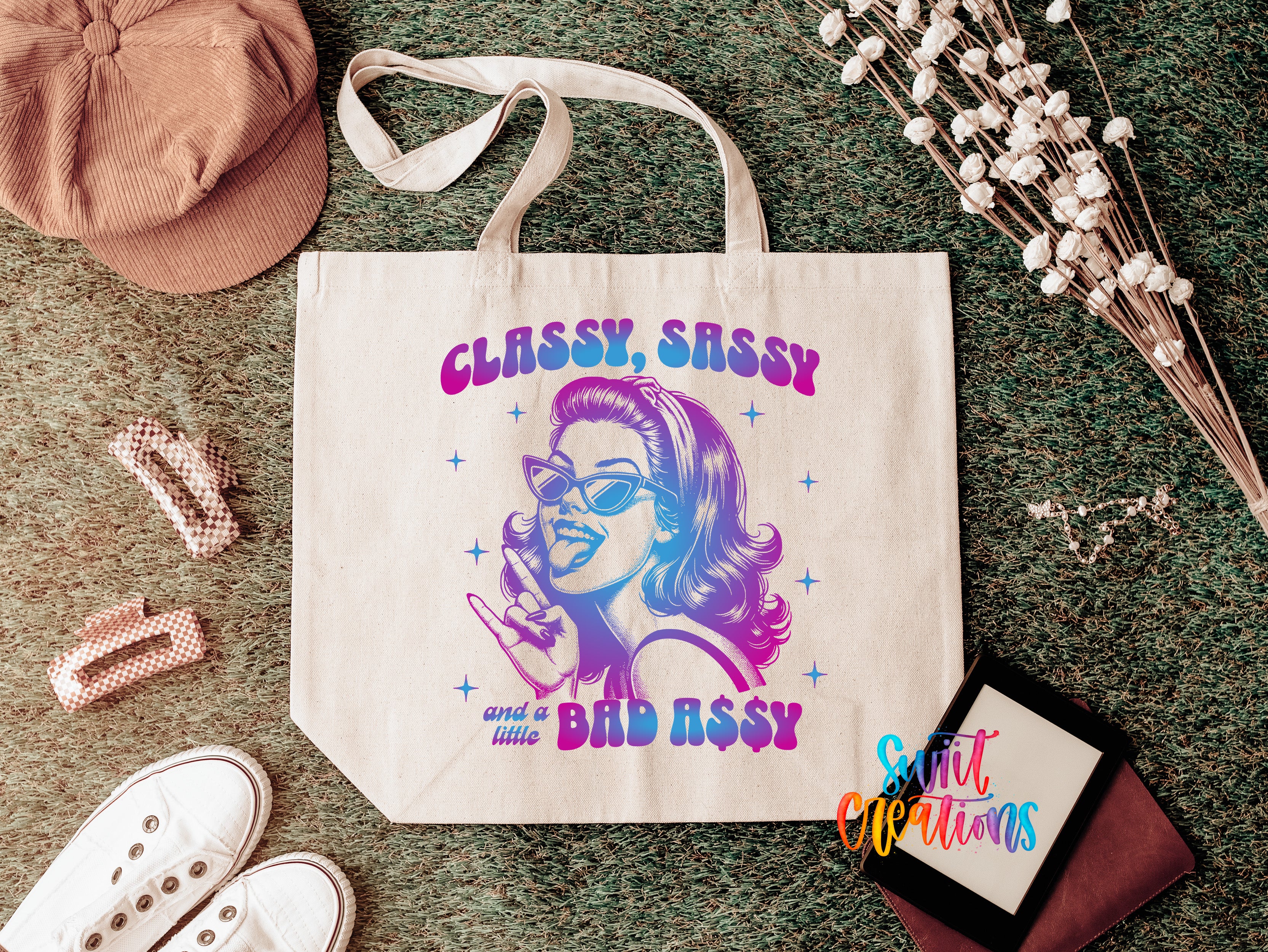 a white tote bag with a colorful graphic design, surrounded by various accessories such as a hat, a pair of shoes, and some flowers.