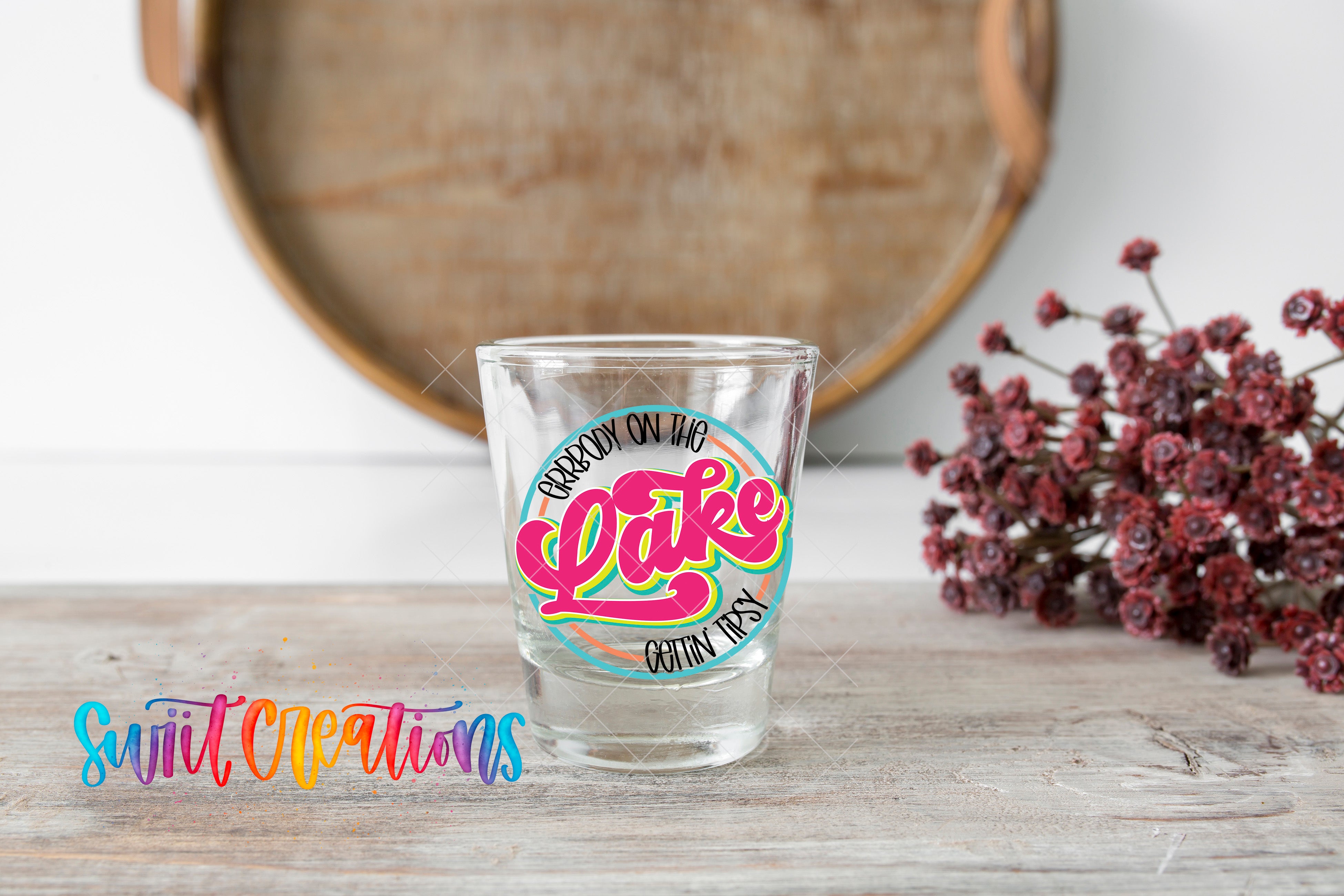 A shot glass with a colorful label that says "BOTTLE ON THE LAKE" is placed on a wooden surface, with a bunch of red flowers in the background.