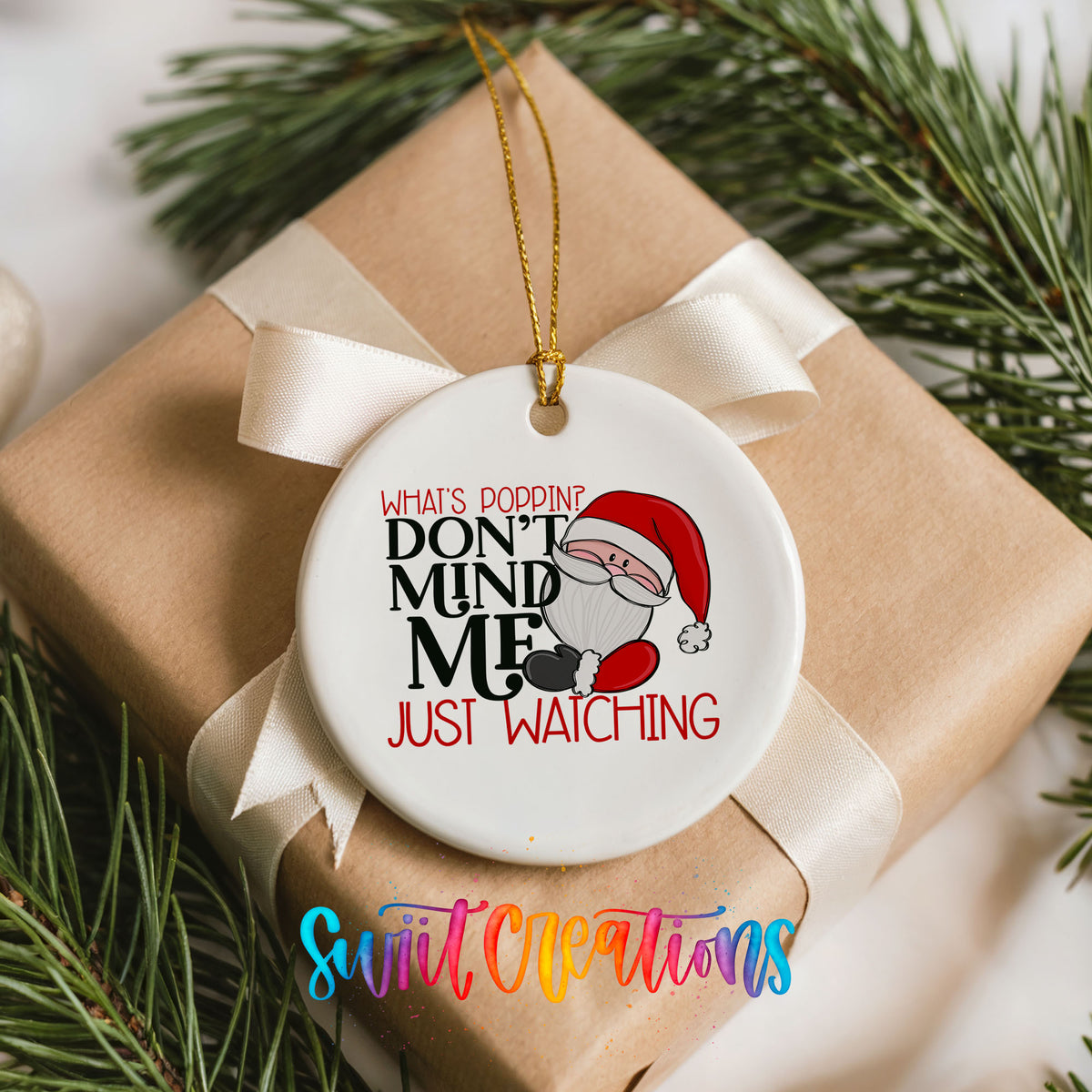 A white ceramic ornament with a Santa Claus design and text that reads "What's poppine don't mind me, just watching". The ornament is hanging from a ribbon and is placed on top of a brown gift box, which is surrounded by pine branches.