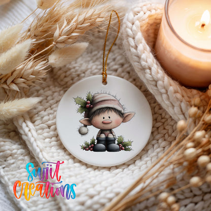 A white ornament featuring a cartoon elf sitting on a branch, surrounded by holly leaves and berries, is displayed on a white knitted surface.