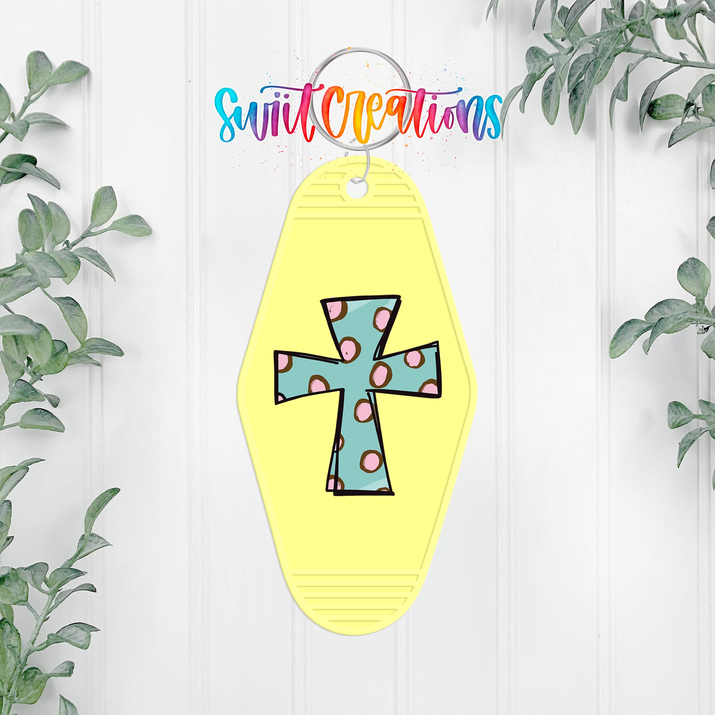 A yellow keychain with a colorful cross design is hanging on a white wall, surrounded by greenery.