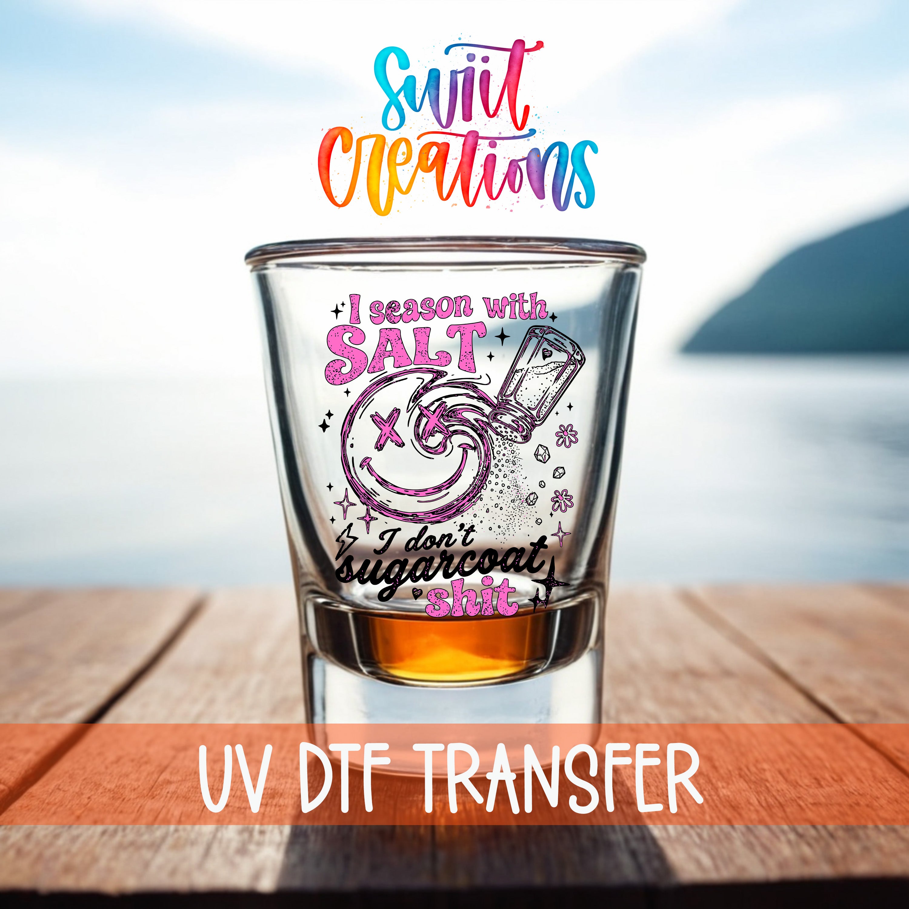 A shot glass with a colorful design and text, placed on a wooden surface with a body of water in the background.