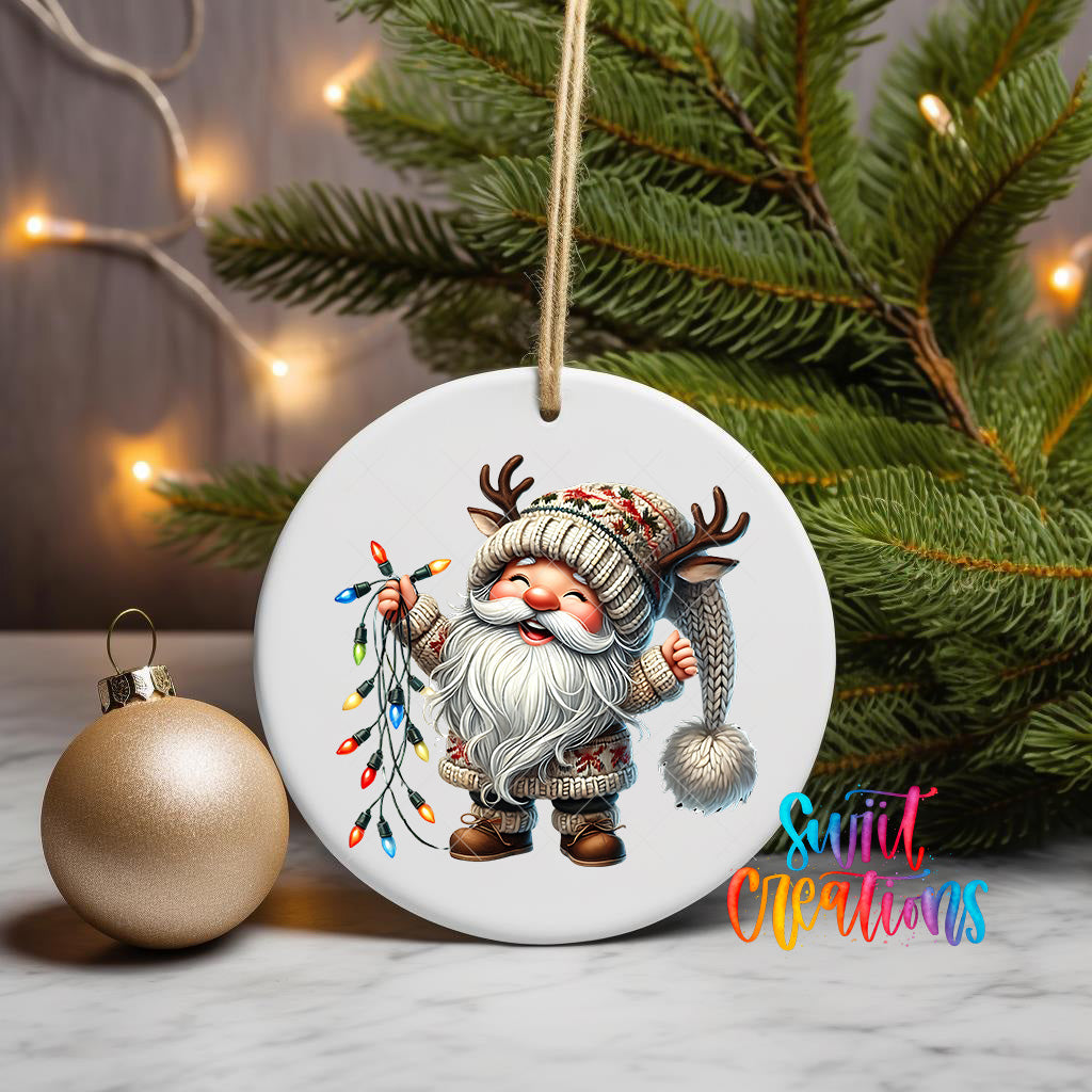 A white ornament featuring a smiling gnome holding a string of colorful Christmas lights, with a Christmas tree and twinkling lights in the background.