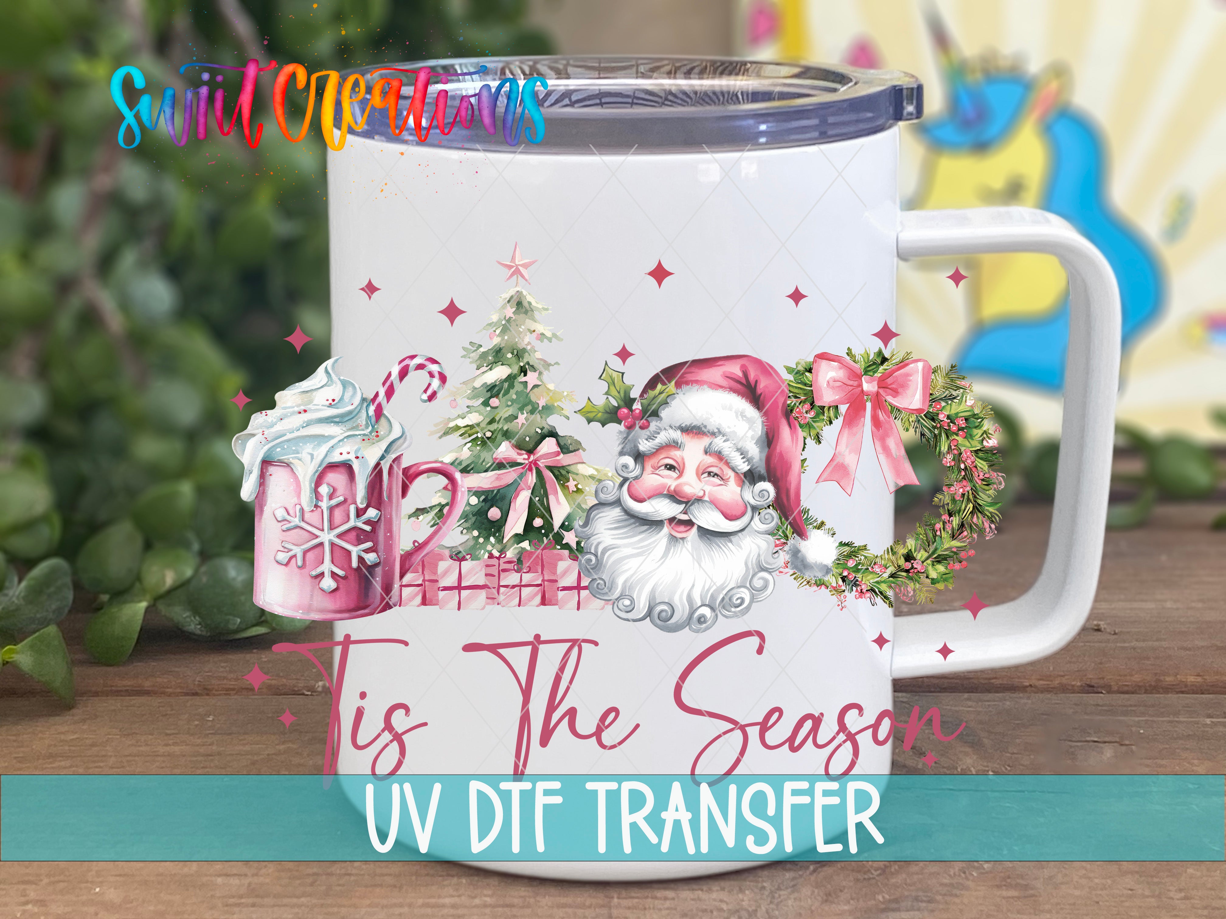 A white mug with a Christmas-themed design, including a Santa Claus face, snowflakes, and a wreath. The mug is placed on a wooden surface, and there is a blurred background that appears to be a garden or outdoor setting.