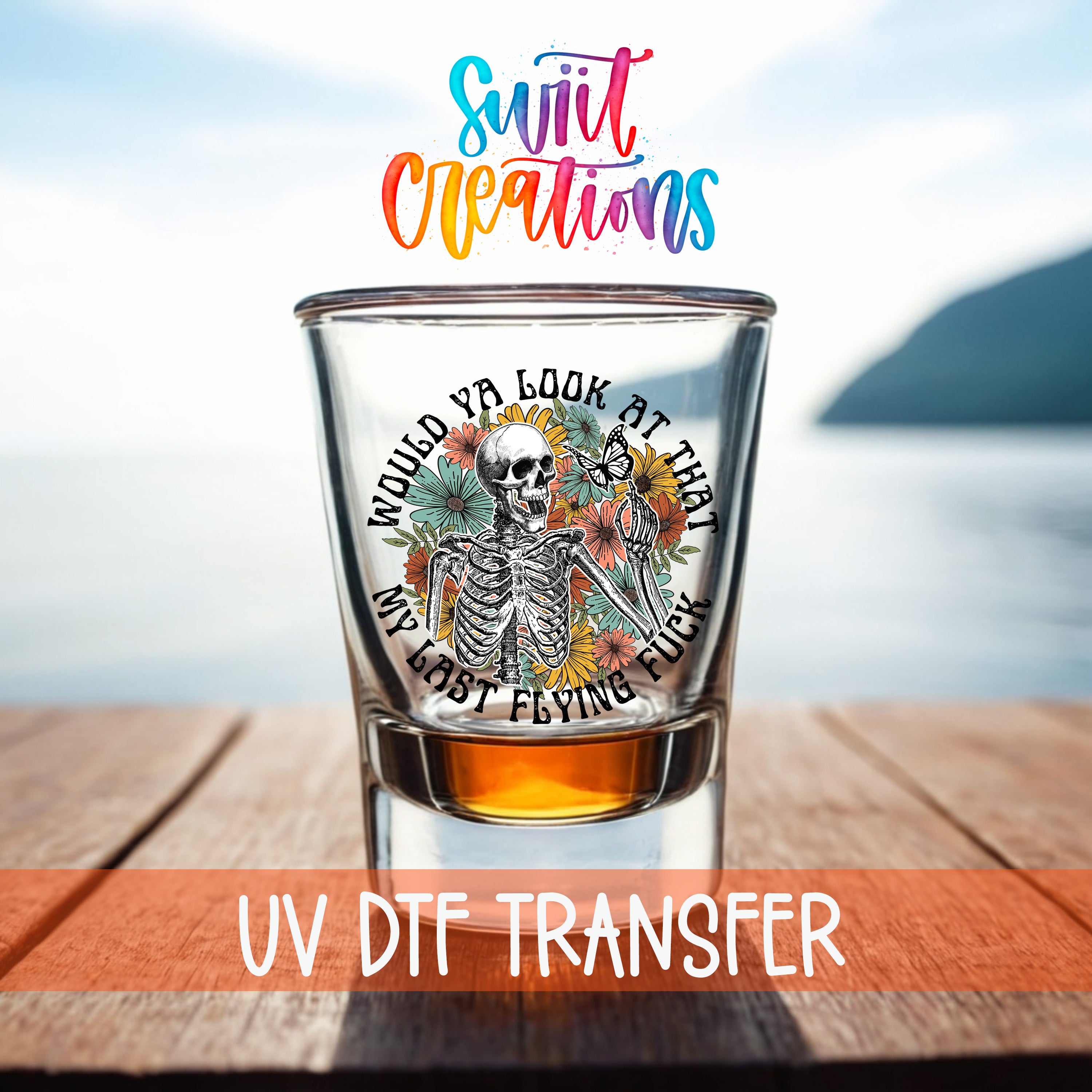 A glass with a skeleton design and text, placed on a wooden surface with a body of water in the background.