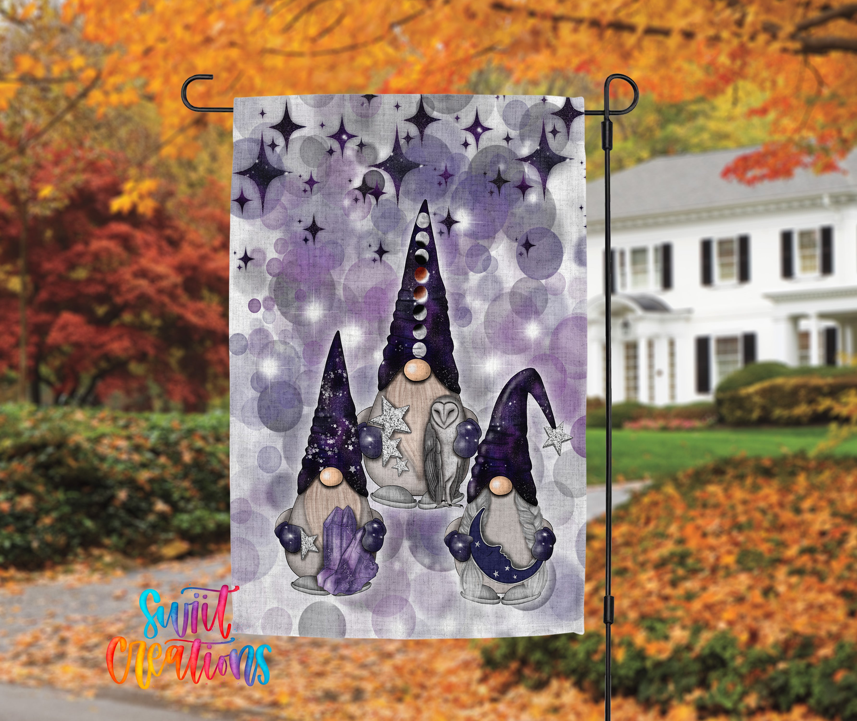 A garden flag featuring three gnomes with purple hats and star-shaped accessories, set against a backdrop of autumn foliage and a white house.
