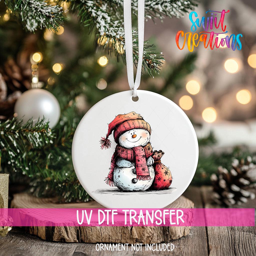 A white ornament with a snowman design hangs on a Christmas tree, surrounded by pine branches and twinkling lights.