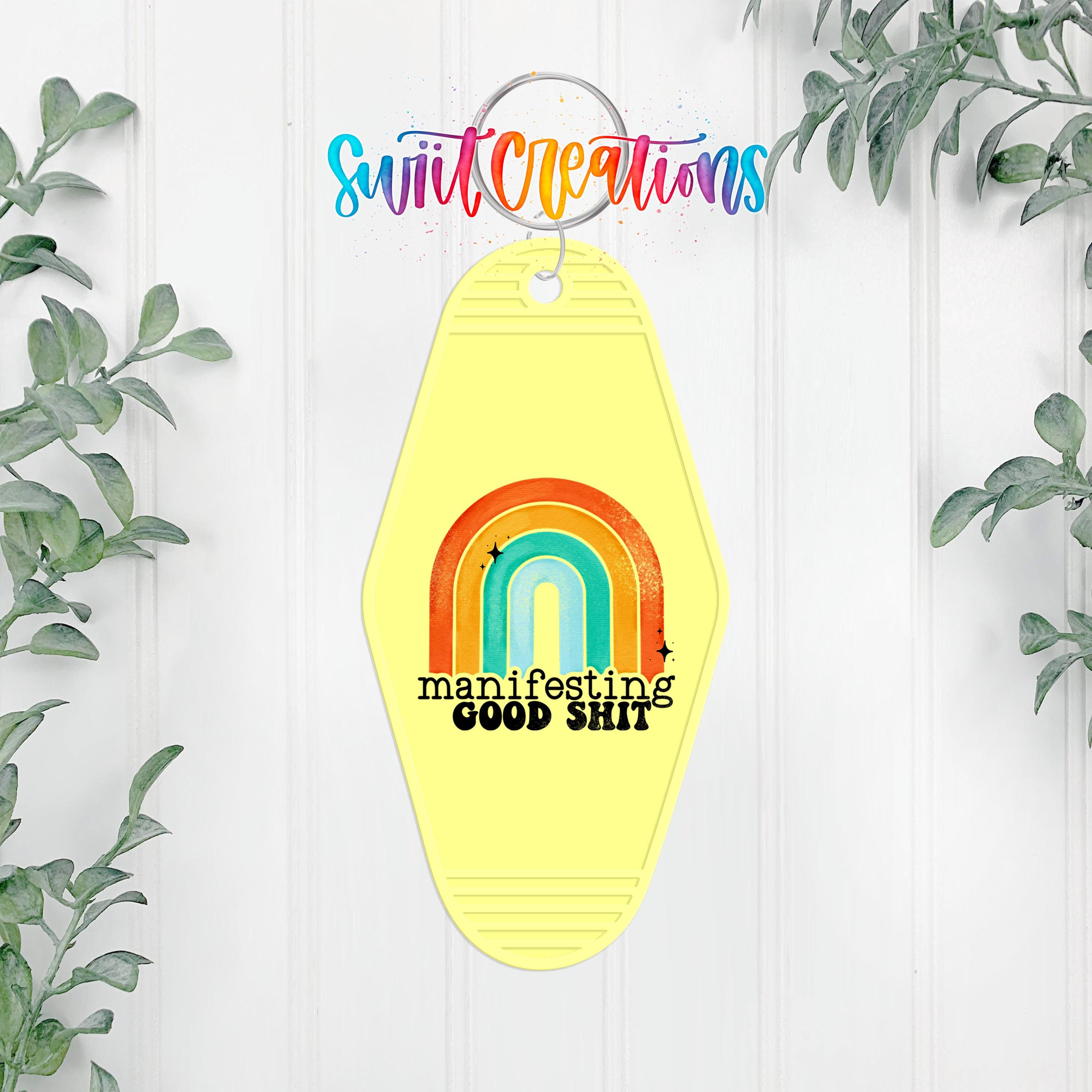 A yellow keychain with a rainbow graphic and the text "manipulating good shit" hanging on a wall, surrounded by greenery.