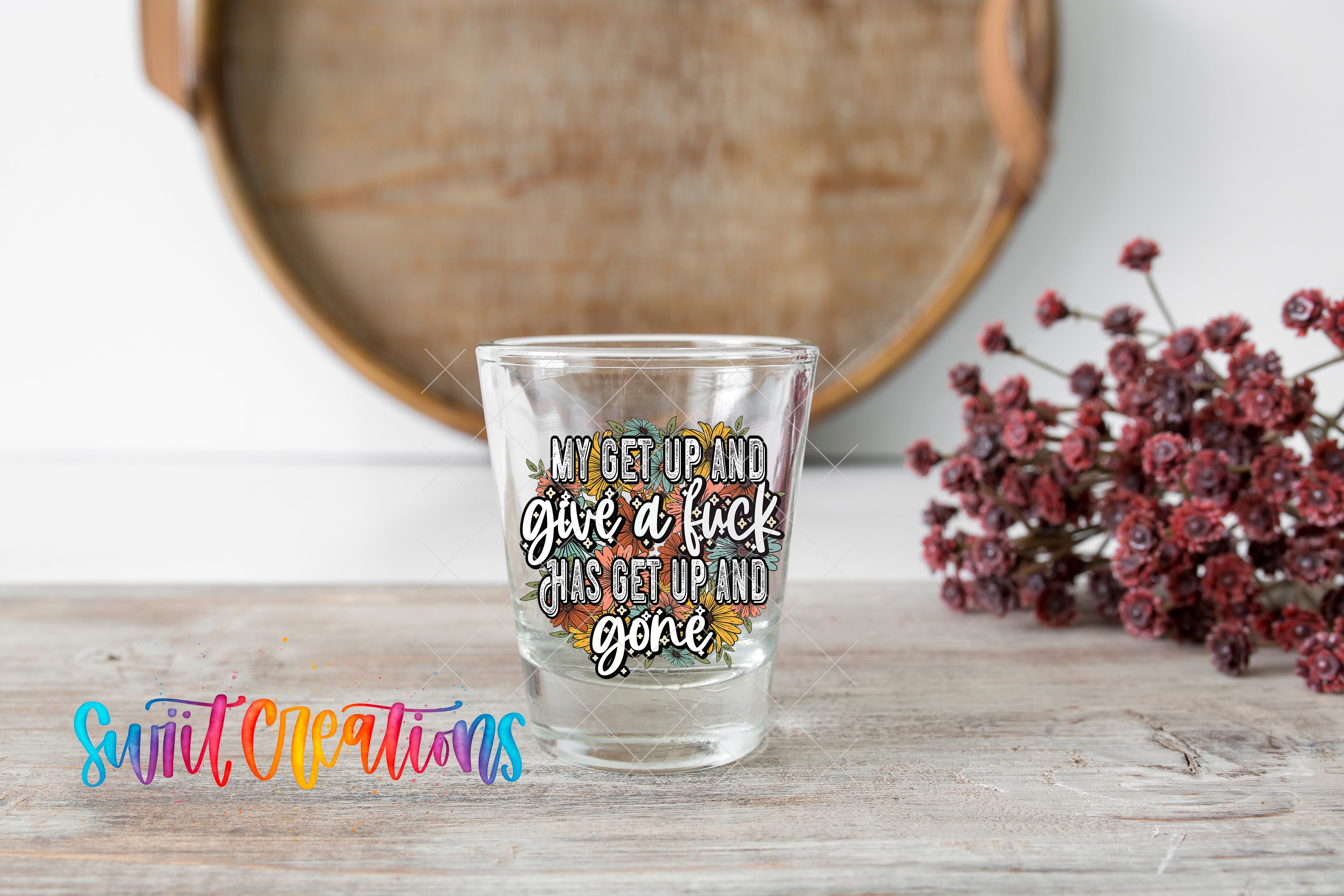 A shot glass with a printed message on it, placed on a wooden surface next to a bunch of red flowers.