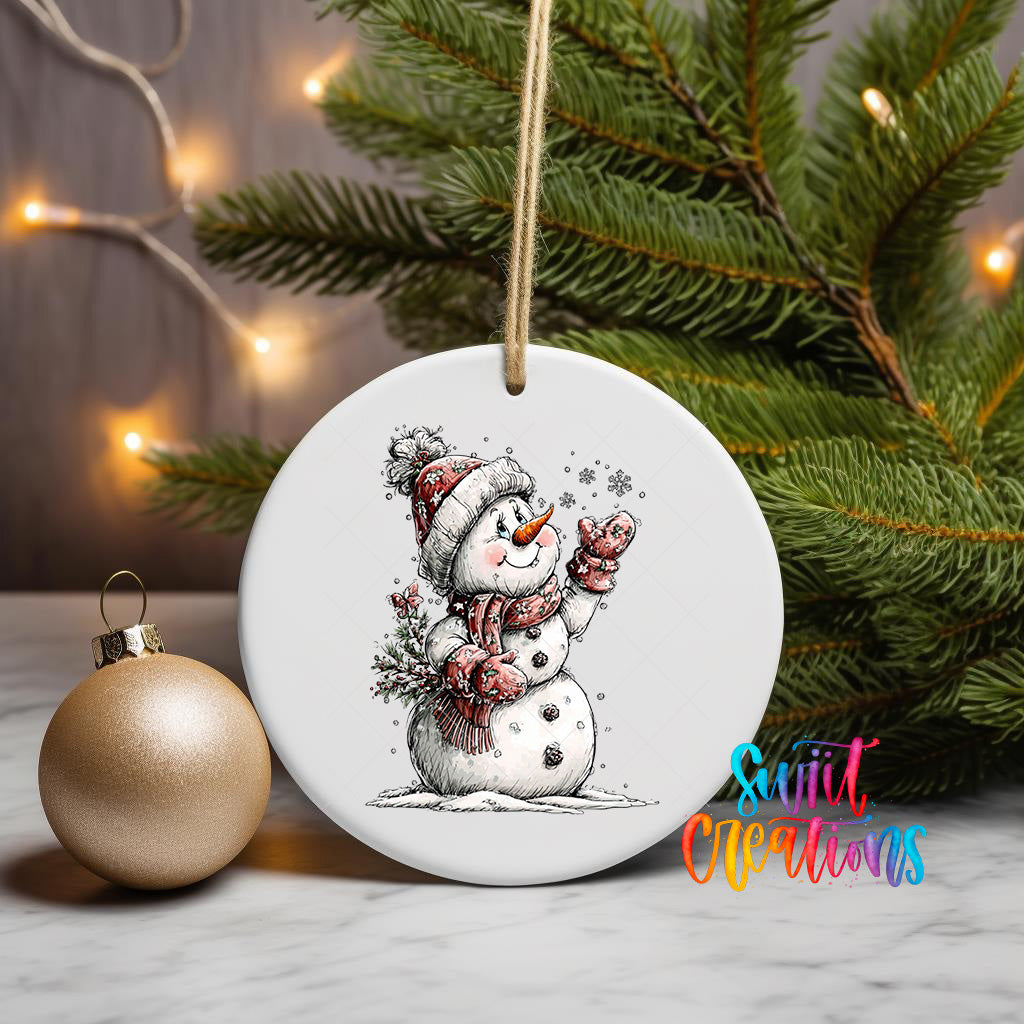 A white ceramic ornament featuring a snowman is displayed on a table, with a gold ornament and a Christmas tree in the background.