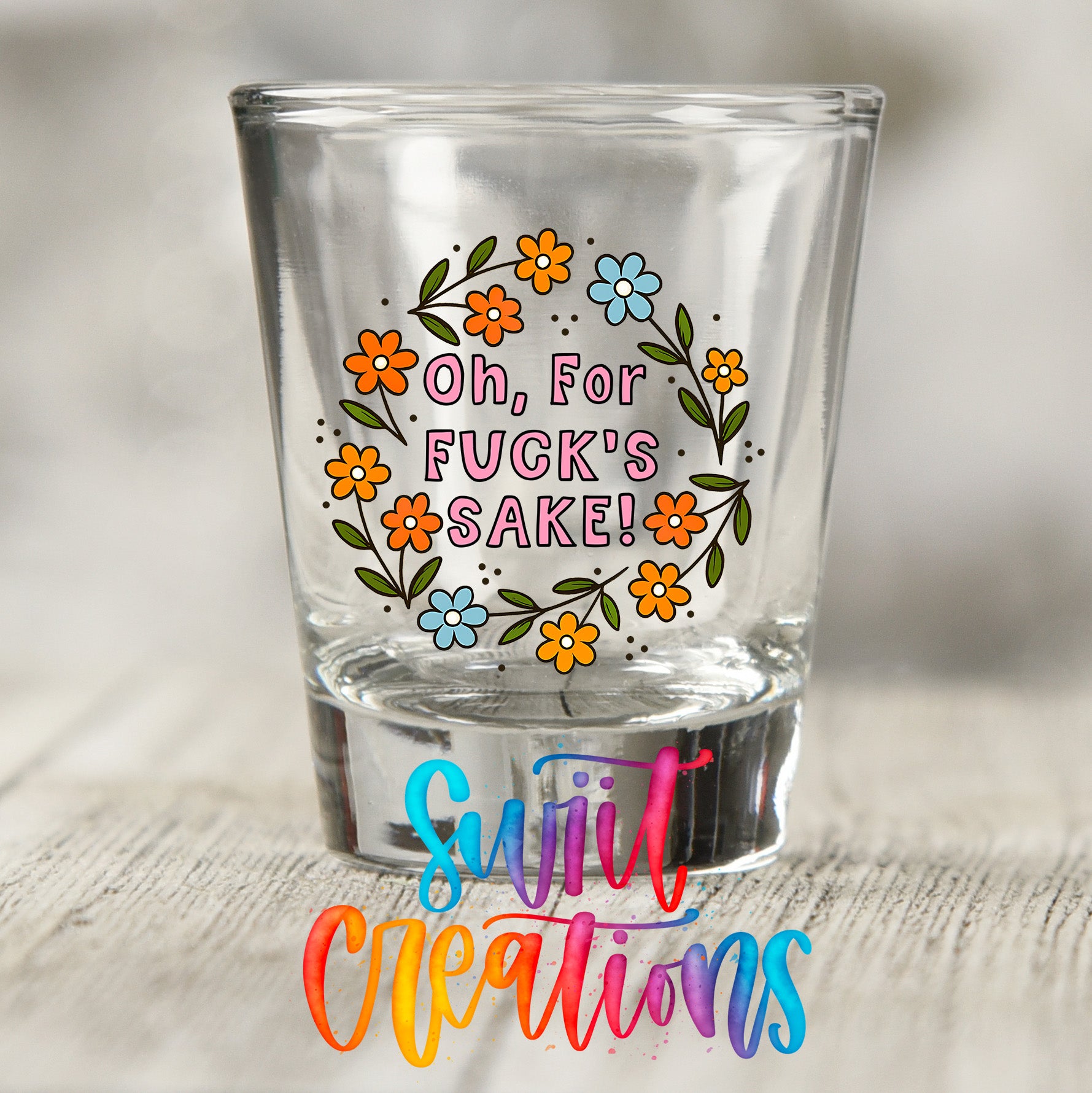 A shot glass with a colorful floral design and text on it, placed on a wooden surface.