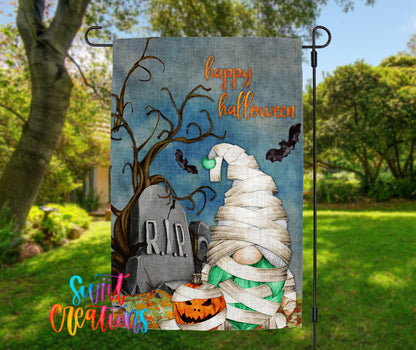 A Halloween-themed garden flag featuring a gnome wrapped in bandages, a carved pumpkin, a tombstone, and bats flying around.