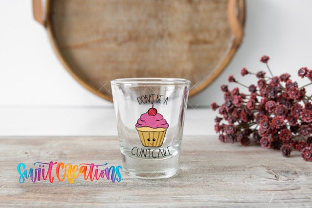 A shot glass with a cupcake design and the text "Don't be a Cuntcake" on it, placed on a wooden surface next to a bunch of red flowers.