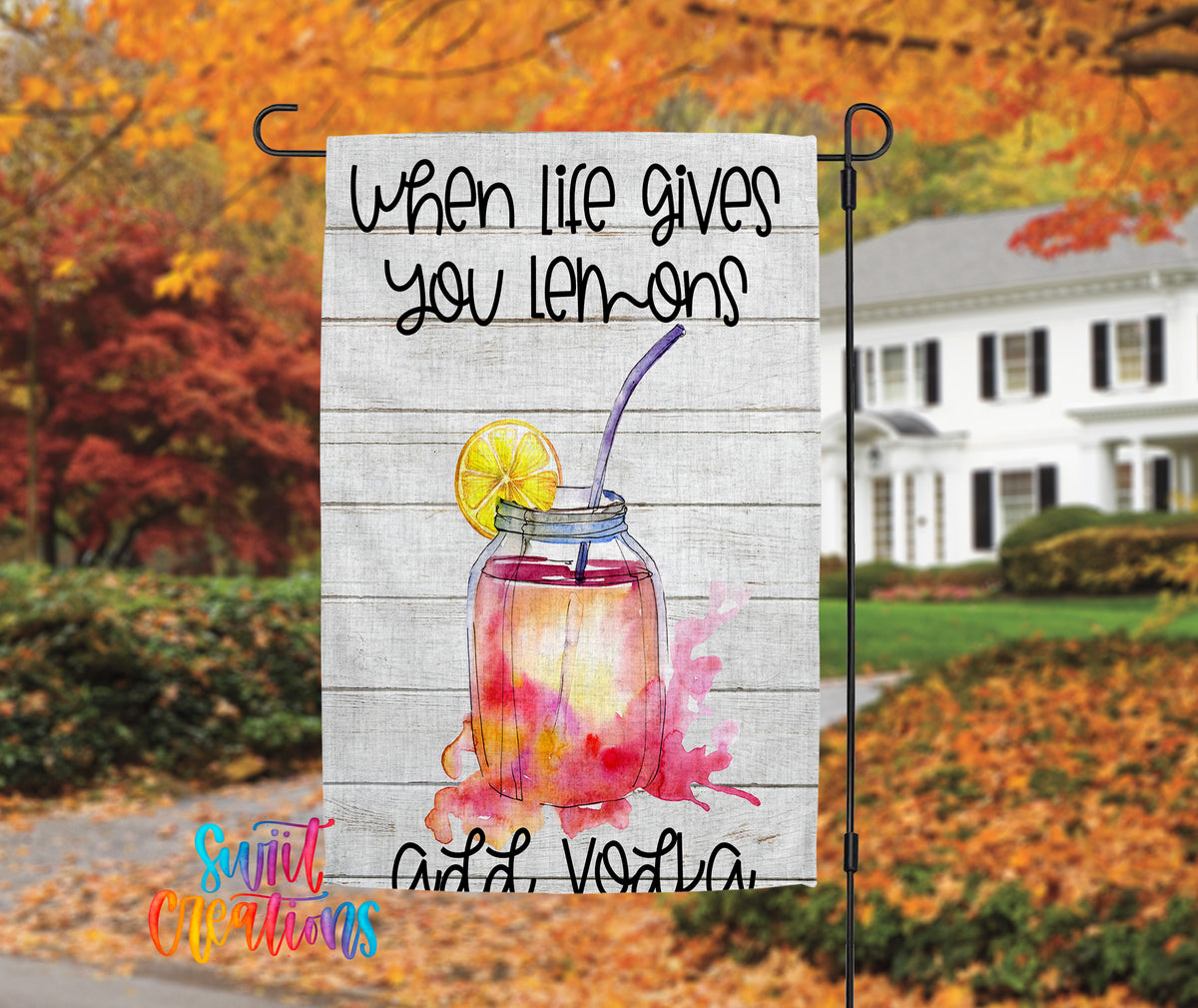 A banner with a lemonade drink and a lemon slice, hanging in front of a house with autumn foliage in the background.