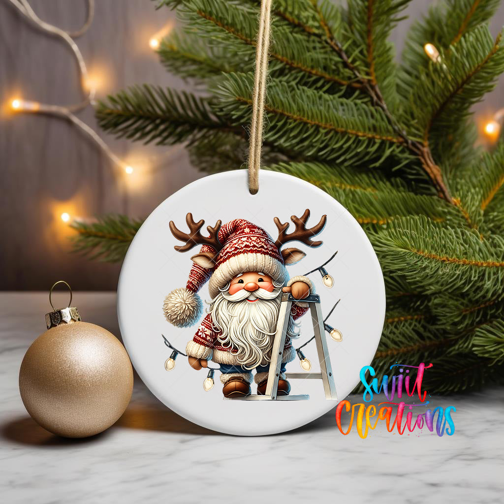 A white ceramic ornament featuring a smiling gnome with antlers, wearing a red and white knitted hat, and holding a ladder, is displayed on a table next to a gold ornament and a Christmas tree.