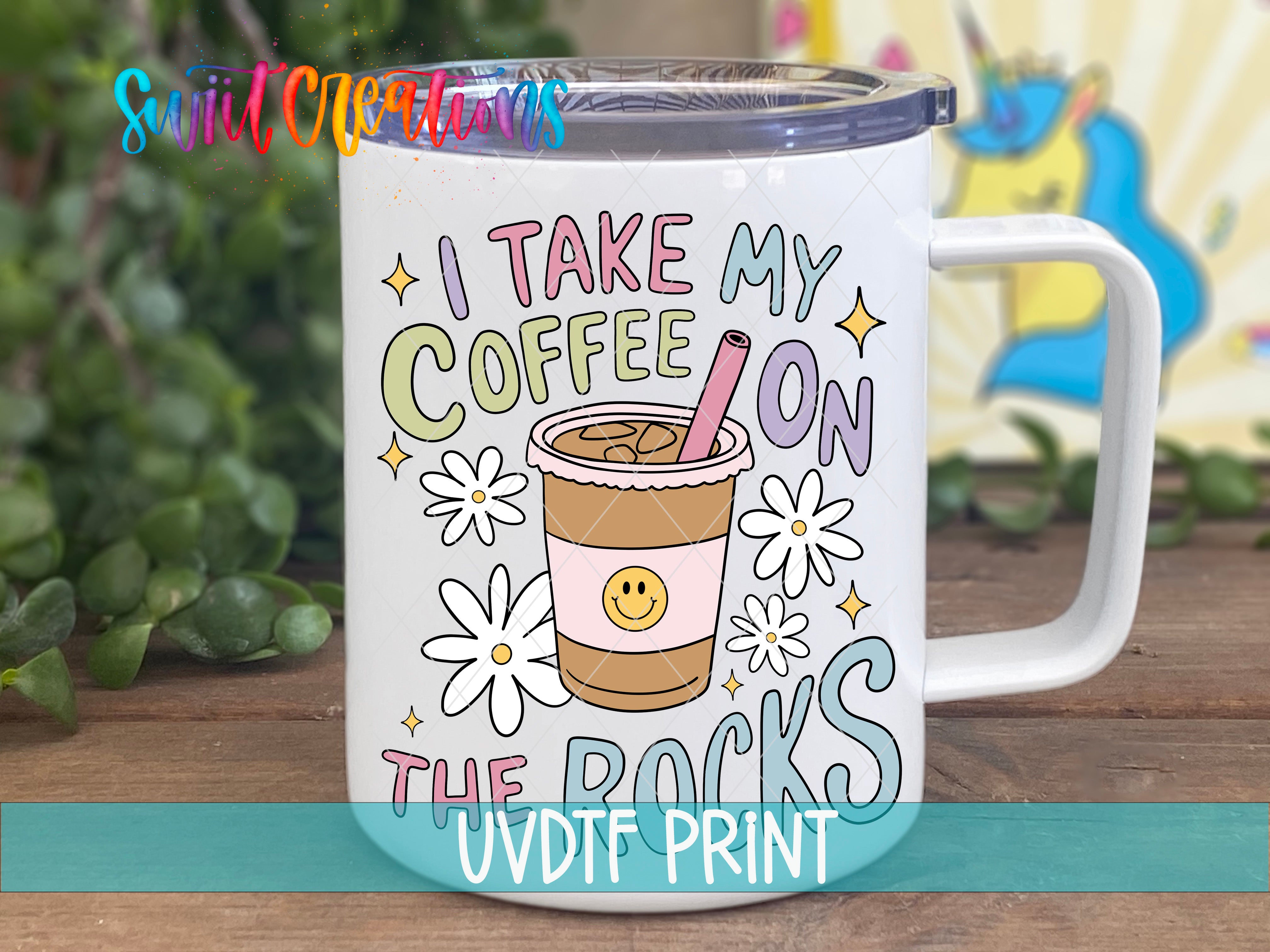 A white mug with a colorful design featuring a coffee cup and flowers, along with the text "I TAKE MY COFFEE ON THE ROCKS" printed on it.
