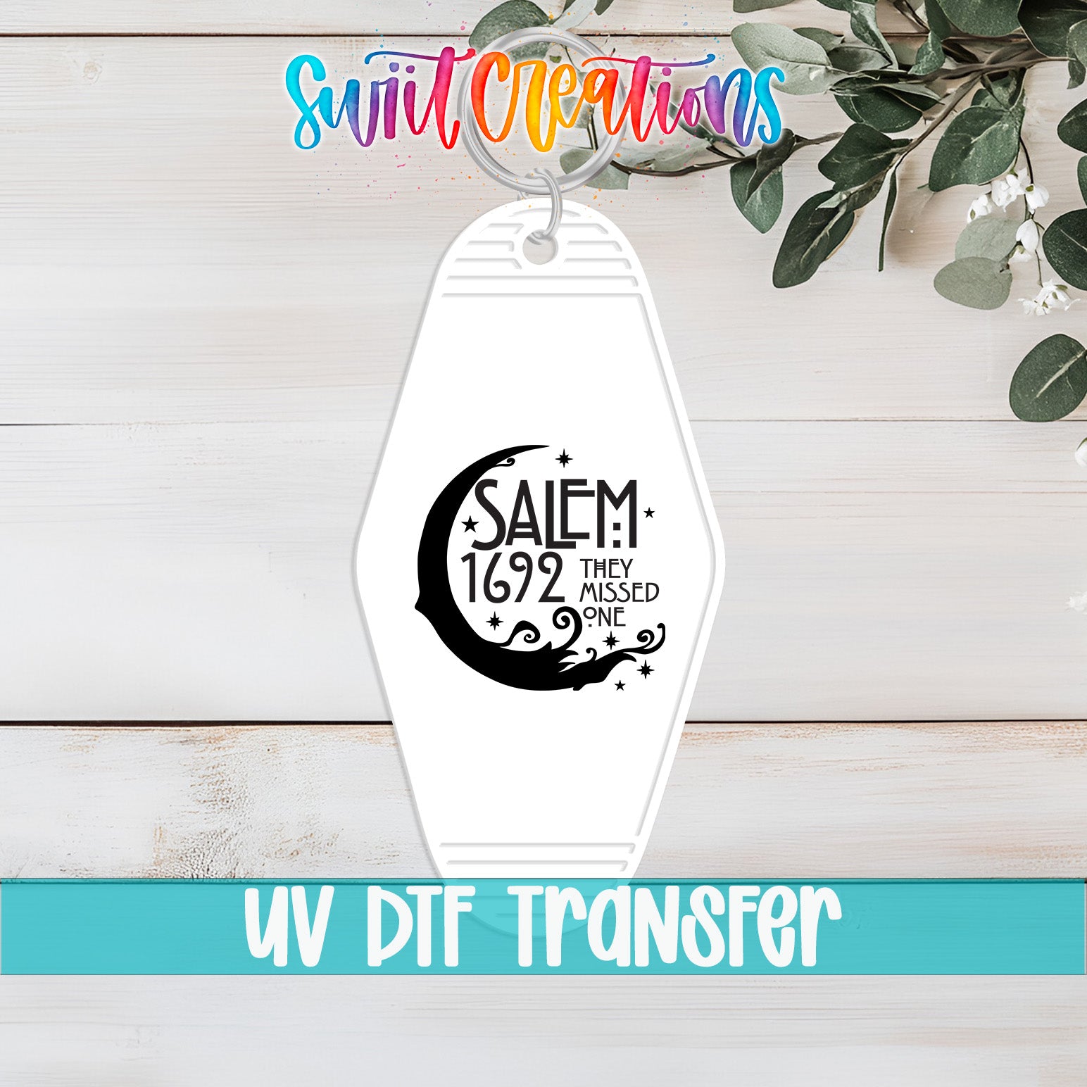 A keychain with a design featuring a crescent moon and stars, along with the text "SAEM 1692 THEY MISSED ONE" printed on it. The keychain is placed on a wooden surface, and there is a plant in the background.