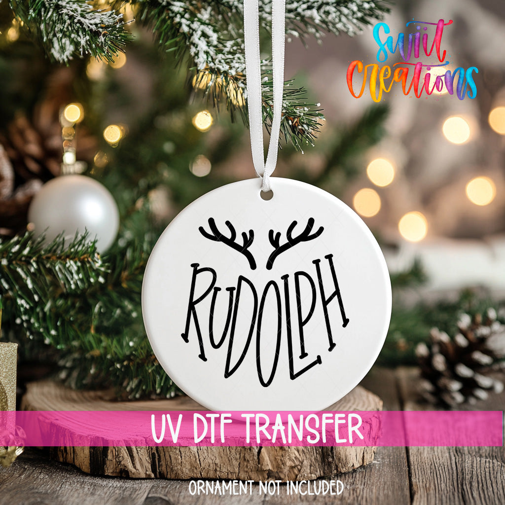 A white ornament with the word "RUDOLPH" written on it, hanging from a Christmas tree.