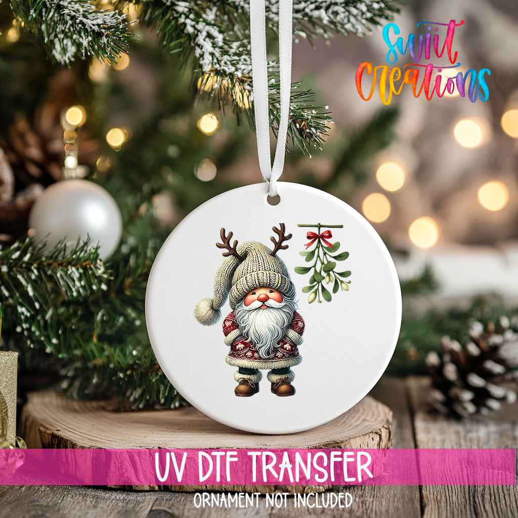 A white ornament with a gnome and mistletoe hangs on a Christmas tree, surrounded by pine branches and twinkling lights.