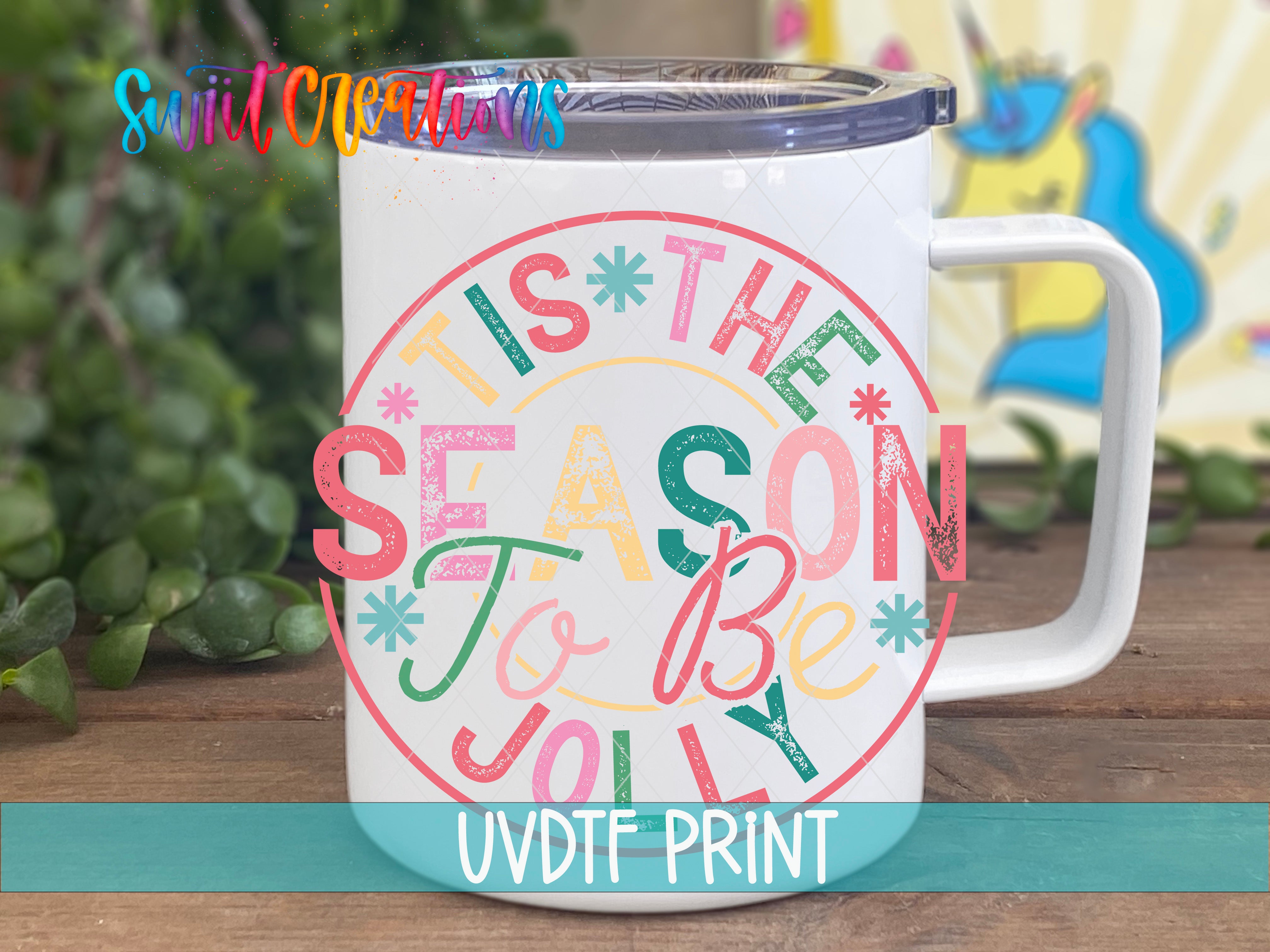 A white mug with a colorful design featuring the text "TIS THE SEASON To Be JOYOUS" in a festive style.