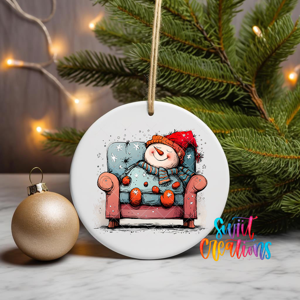 A white ceramic ornament featuring a snowman sitting in a red armchair, with a Christmas tree and twinkling lights in the background.