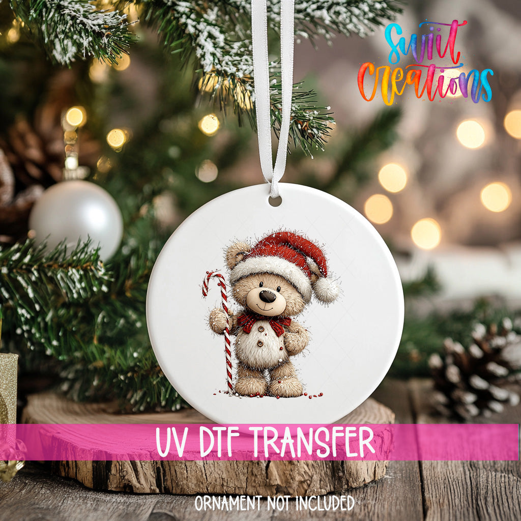 A white ornament with a teddy bear wearing a Santa hat and holding a candy cane is hanging on a Christmas tree.