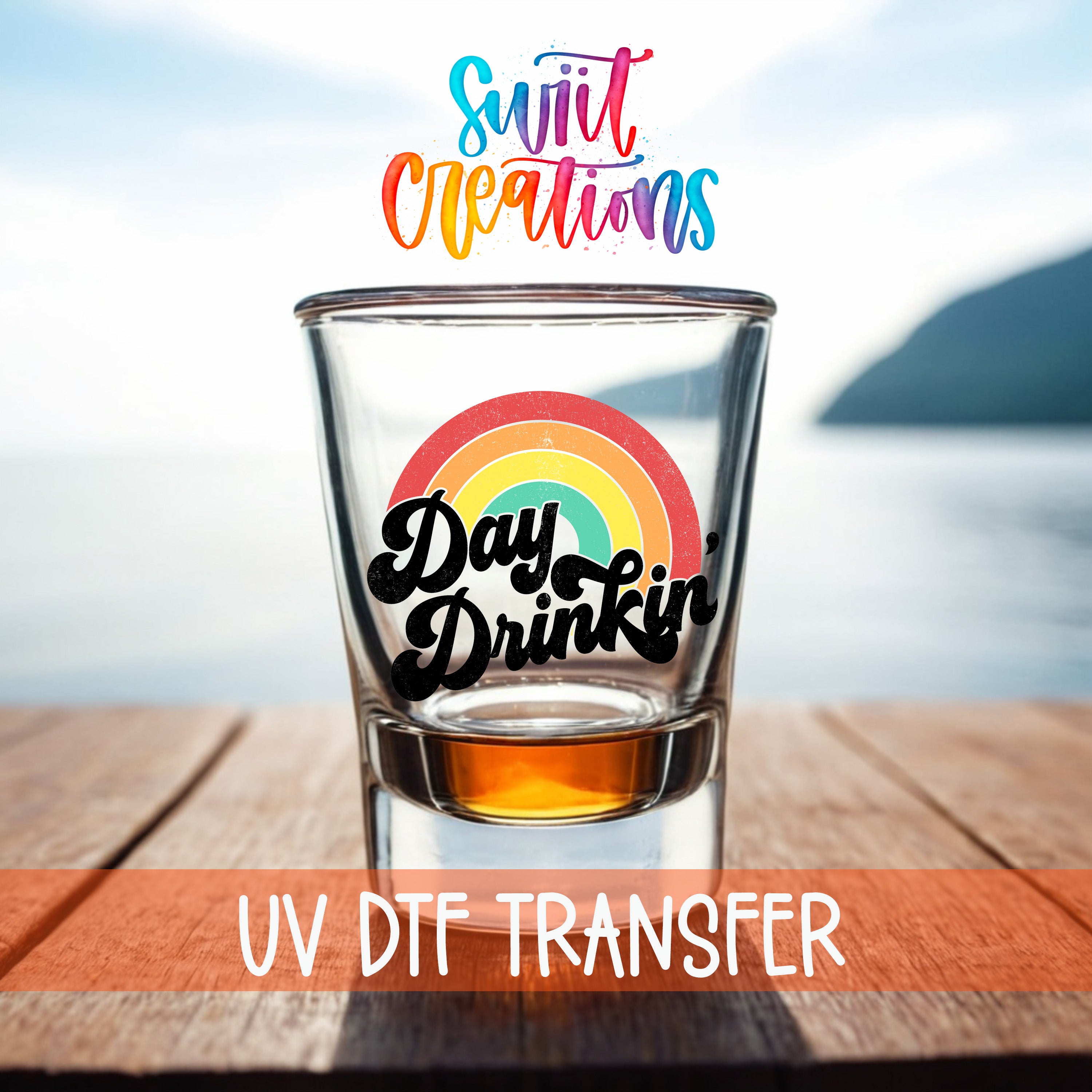 A shot glass with a rainbow design and the text "Day Drinking" on it, placed on a wooden surface with a body of water in the background.