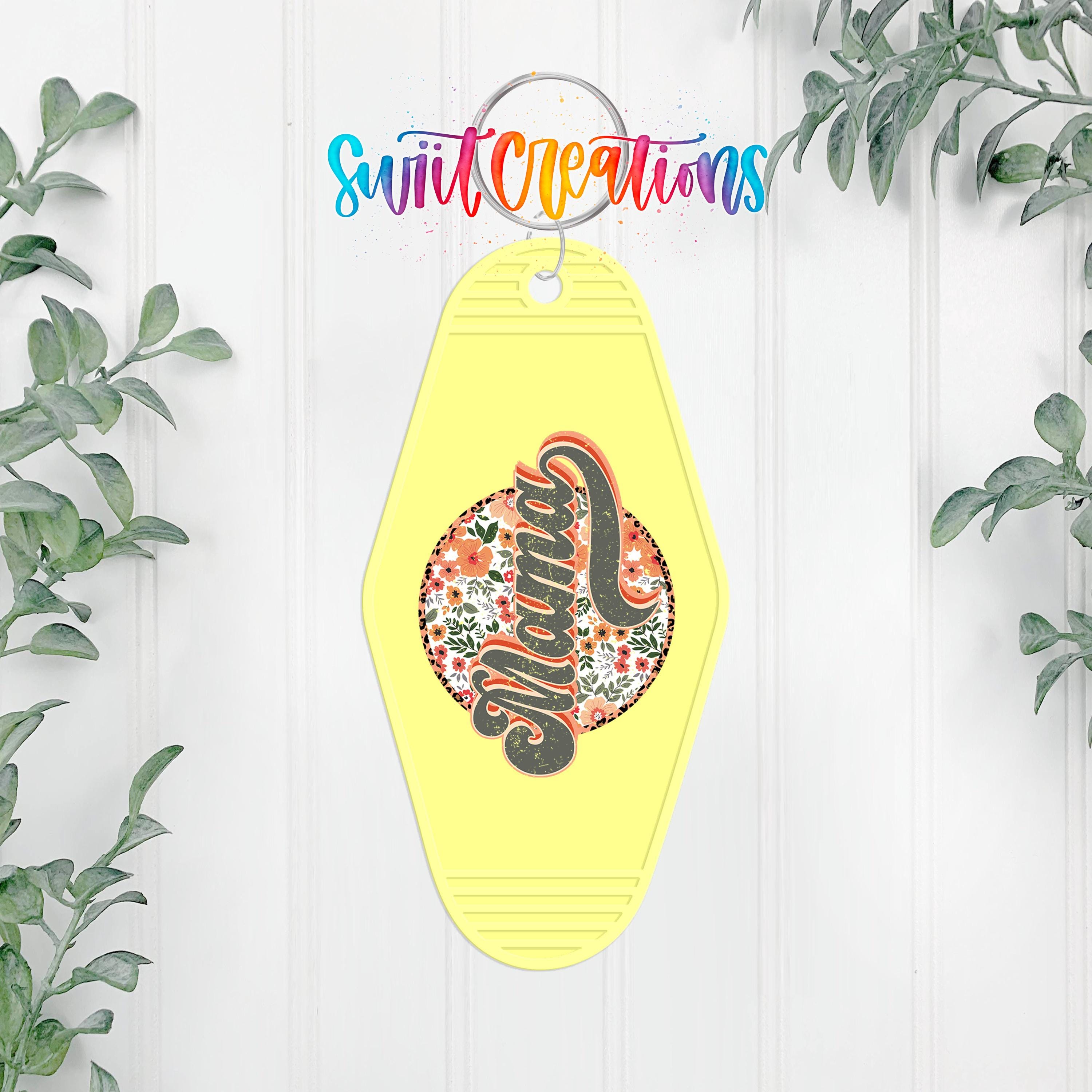 A yellow keychain with a floral design and the word "Mama" written on it, hanging on a white wall with green plants on either side.