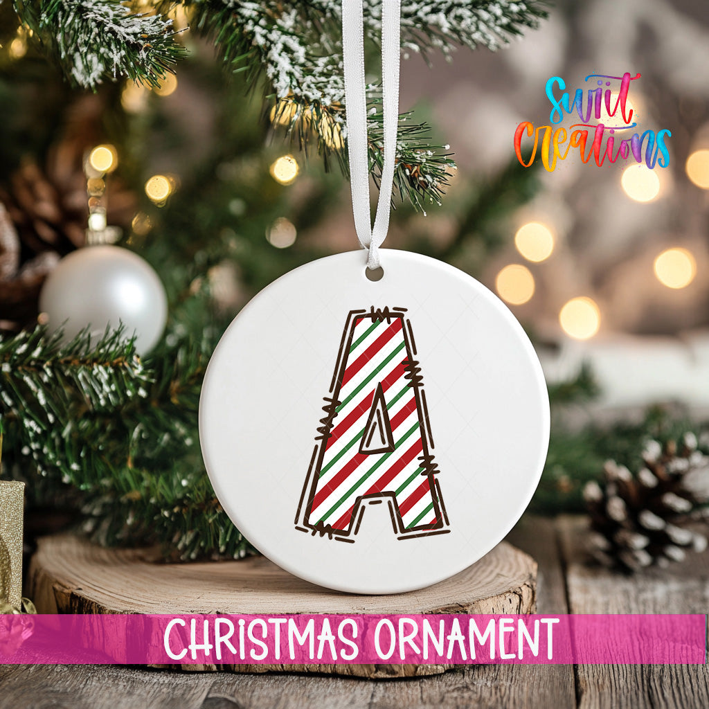 A white Christmas ornament with a red and green striped design hangs from a tree branch, with a blurred background of lights and greenery.