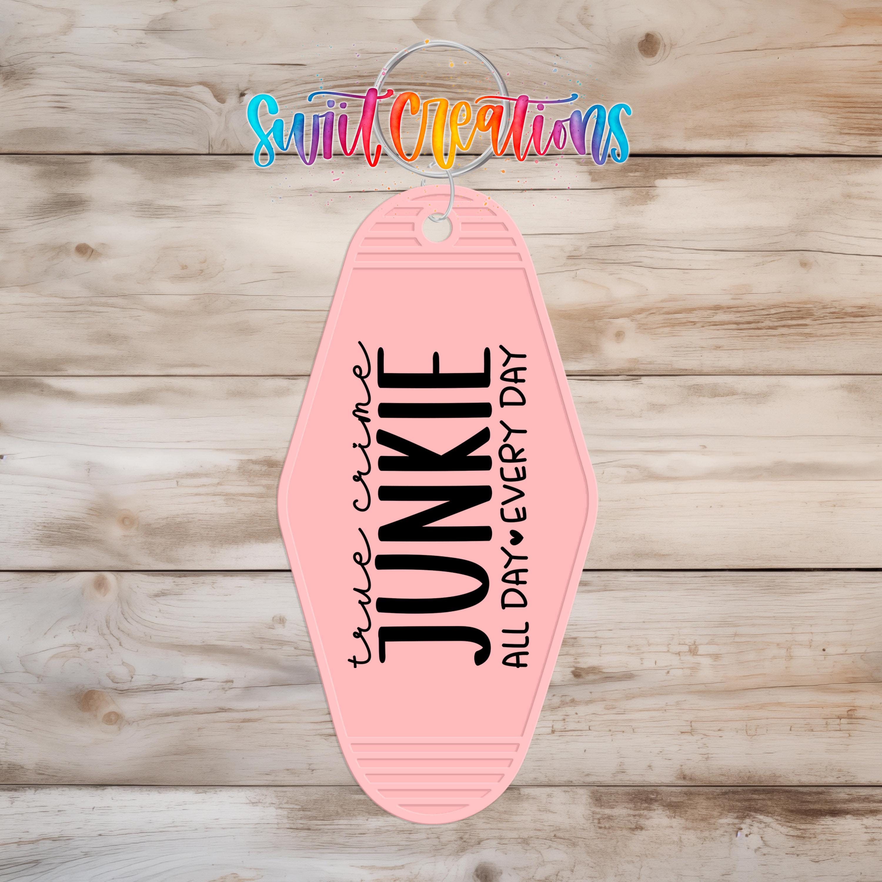A pink keychain with the text "True Crime Junkie All Day Every Day" printed on it, hanging on a wooden surface.
