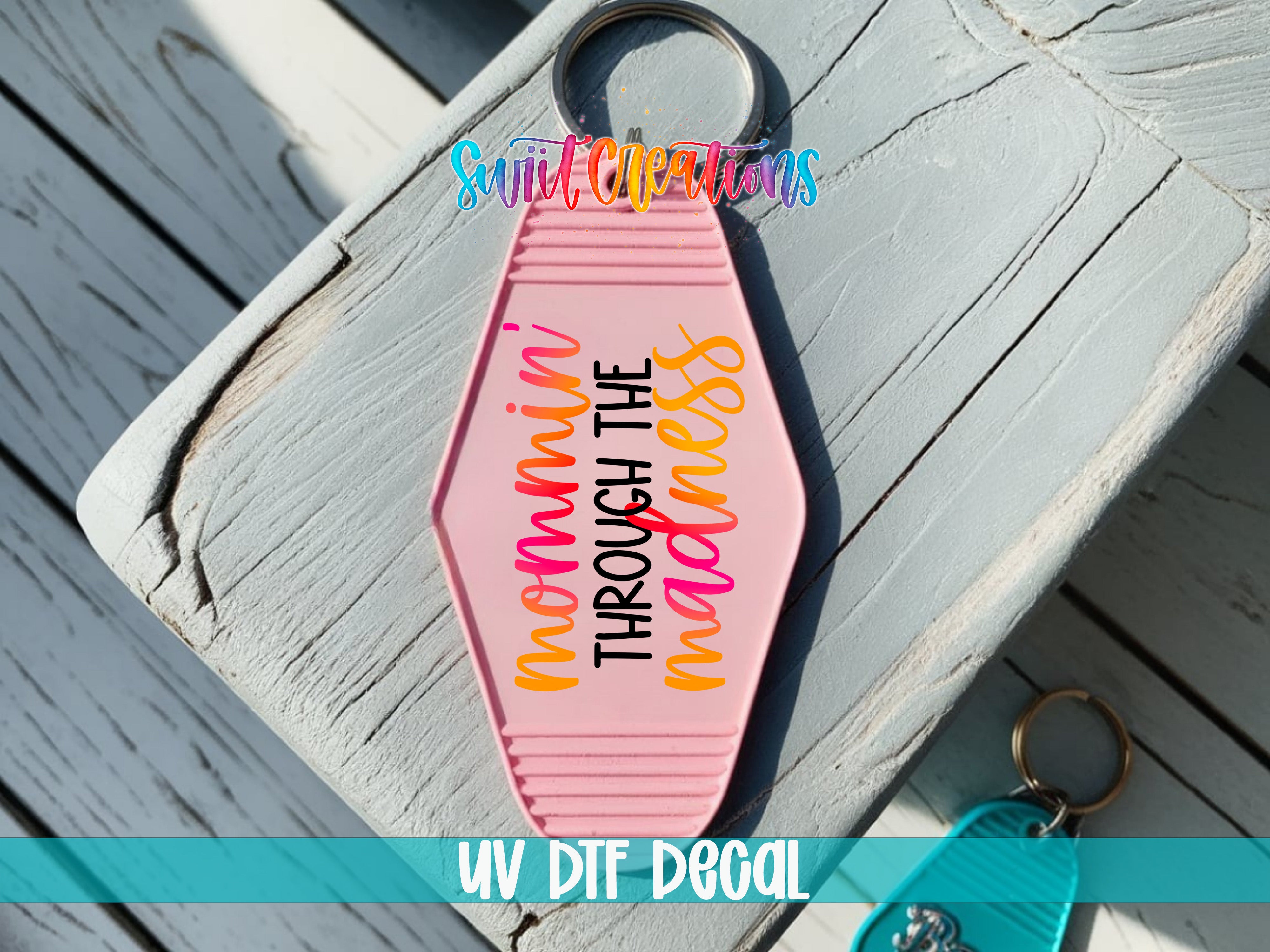 A pink keychain with the text " mommy through the madness" printed on it, placed on a wooden surface.