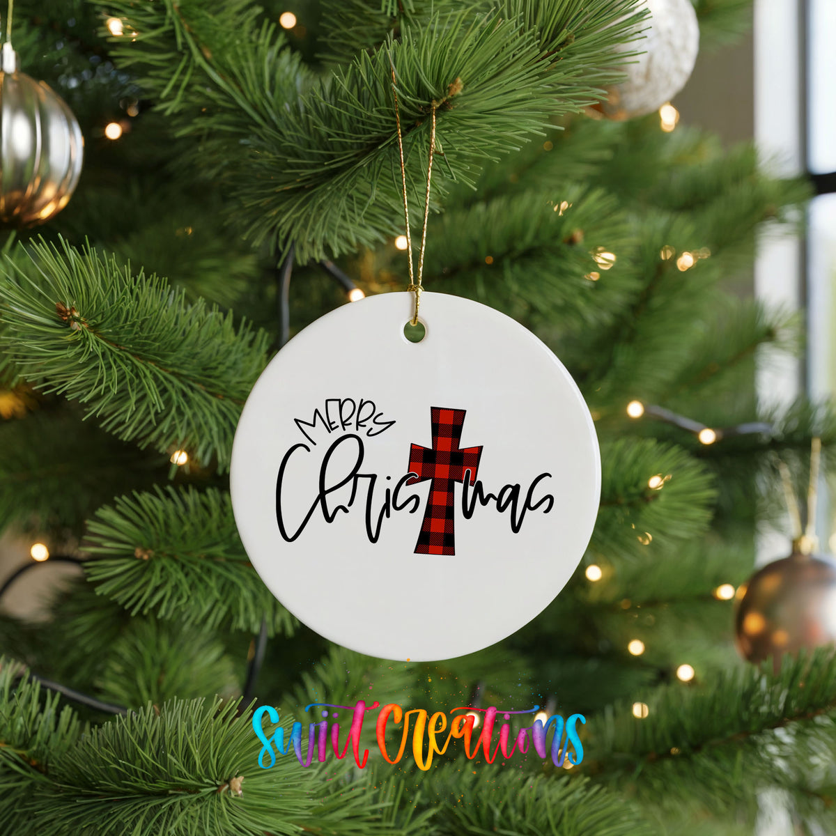 A white Christmas ornament with the text "Merry Christmas" and a red and black plaid cross is hanging on a pine tree.