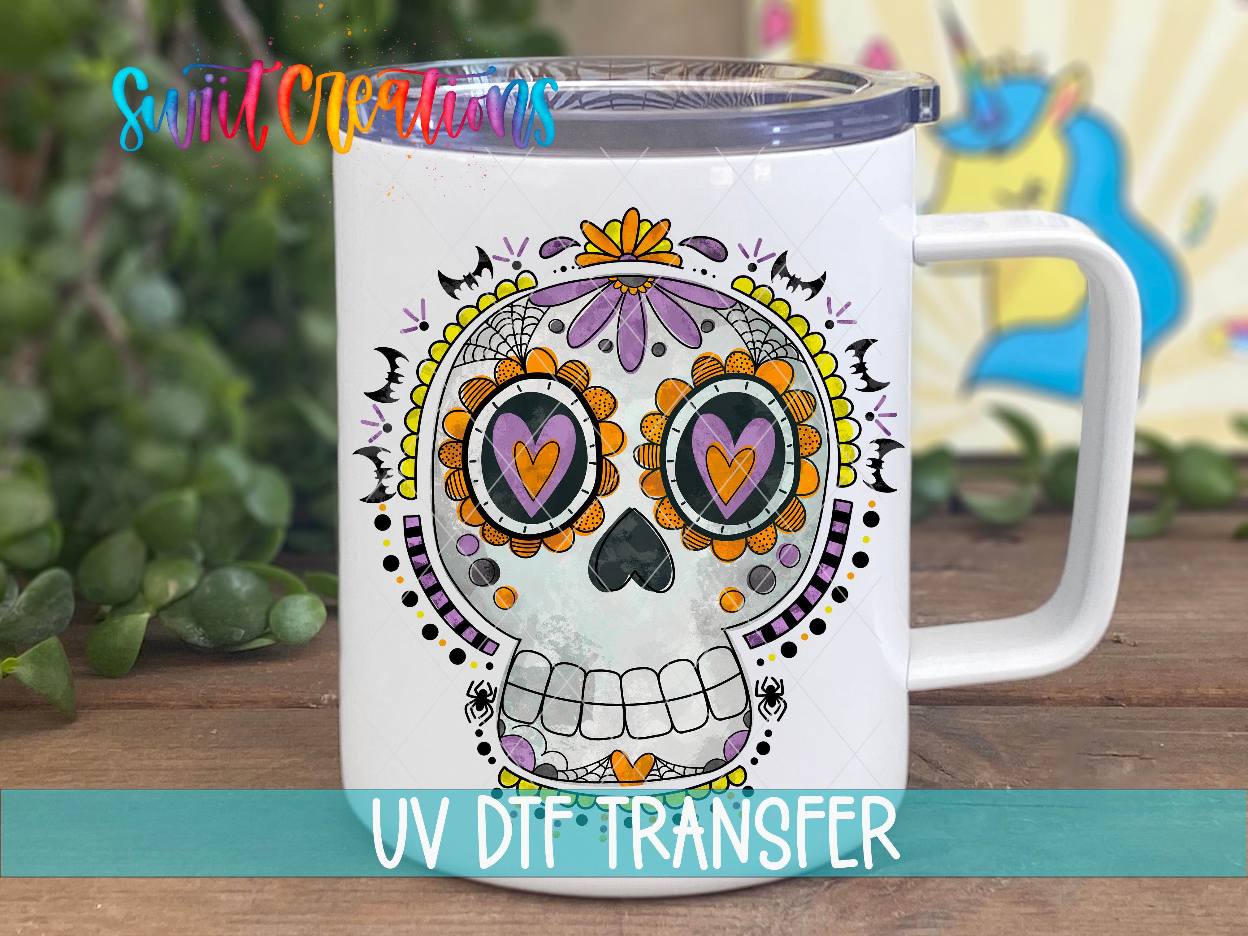 A white ceramic mug with a colorful, intricate skull design featuring a sun-like motif, flowers, and hearts. The mug is placed on a wooden surface, and there is a blurred background that includes some greenery.