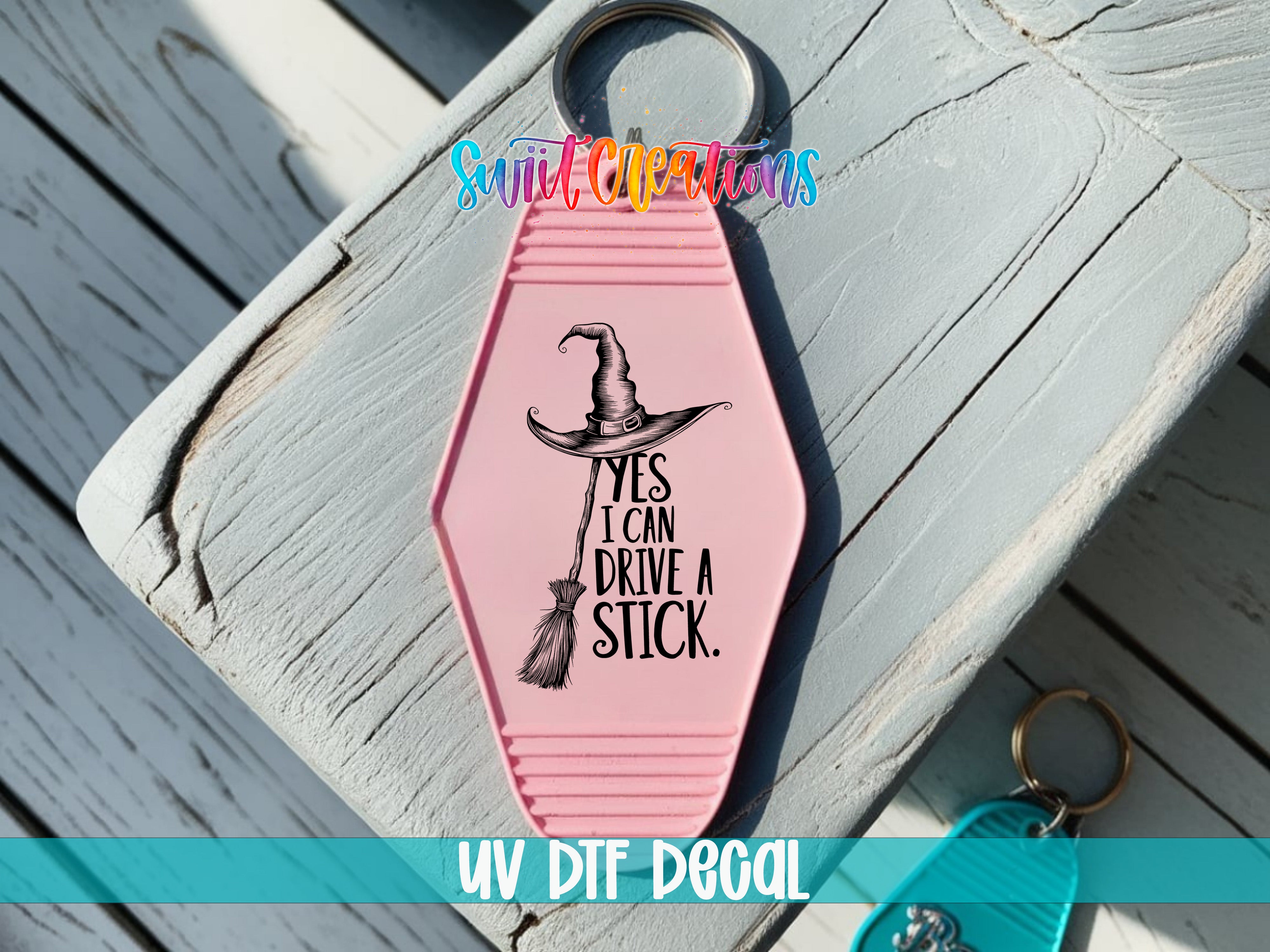 A pink keychain with a witch's hat and the text "YES I CAN DRIVE A STICK" printed on it, placed on a wooden surface.