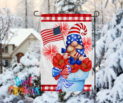 A patriotic-themed garden flag featuring a gnome holding an American flag and surrounded by red and white flowers, set against a snowy winter landscape with a house in the background.