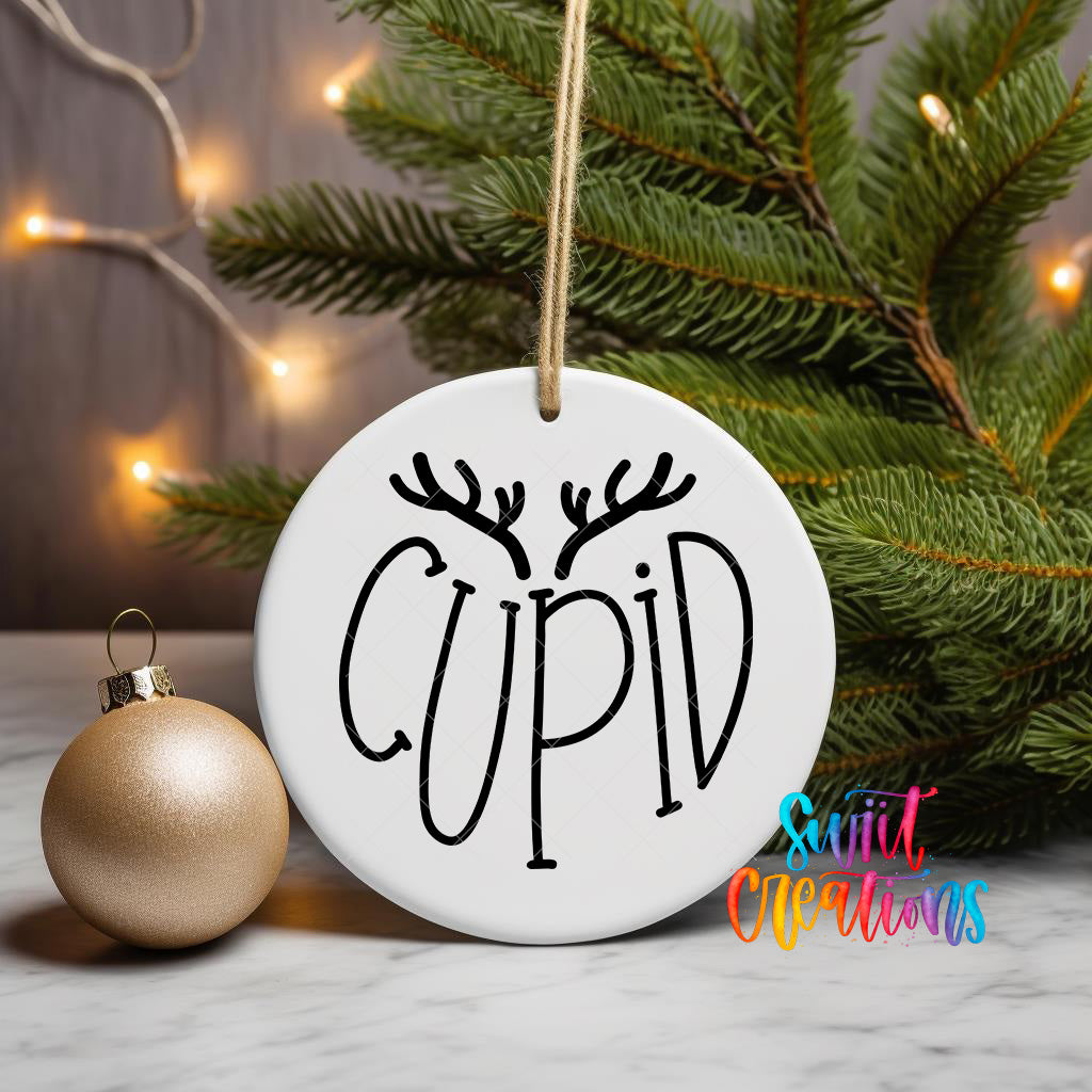 A white ceramic ornament with the word "CUPID" written on it, hanging from a string, is placed next to a gold ornament and a pine tree with twinkling lights in the background.