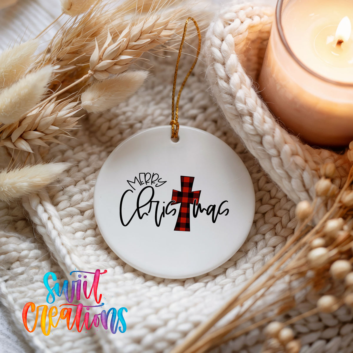 A white ceramic ornament with a red and black plaid cross design, hanging on a white knit blanket. The ornament has the text "Merry Christmas" written on it.