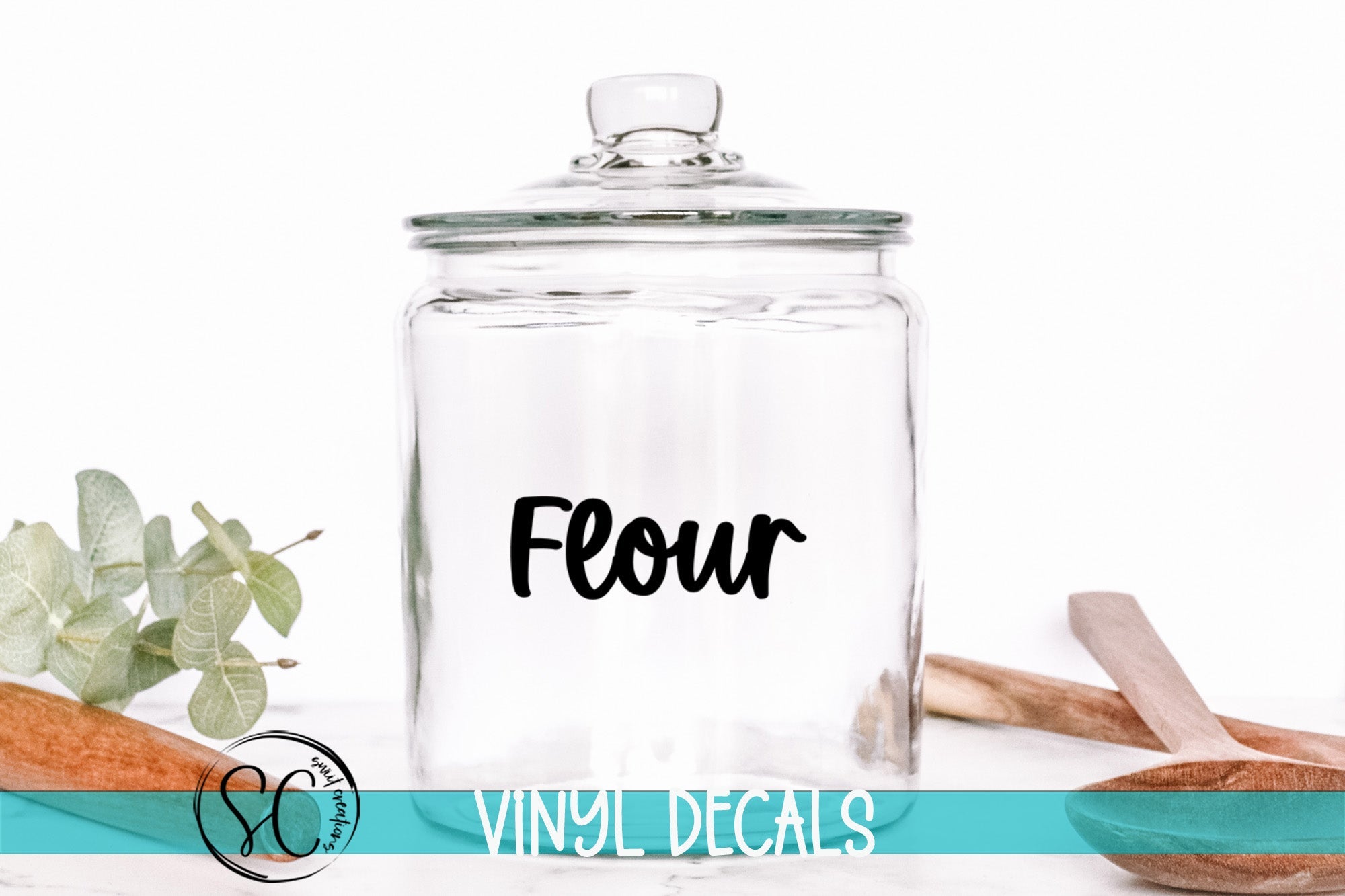 A clear glass jar with a black label that says "Flour" is placed on a white surface, surrounded by various kitchen utensils and decorative items.