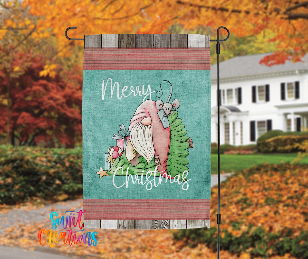A colorful garden flag with a gnome and Christmas decorations, hanging in front of a white house with autumn foliage in the background.