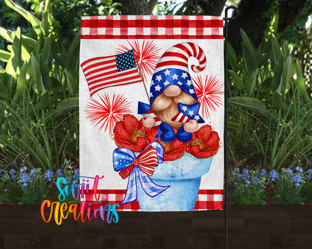A patriotic-themed garden flag featuring a gnome holding an American flag and surrounded by red and white flowers, with a fireworks display in the background.