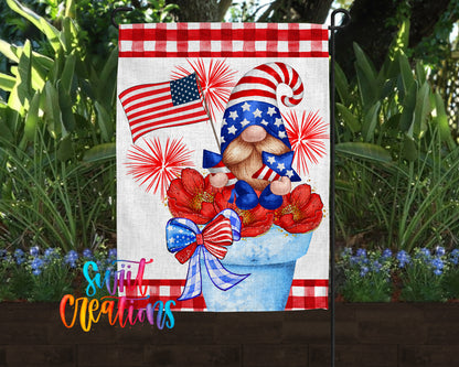 A patriotic-themed garden flag featuring a gnome holding an American flag and surrounded by red and white flowers, with a fireworks display in the background.