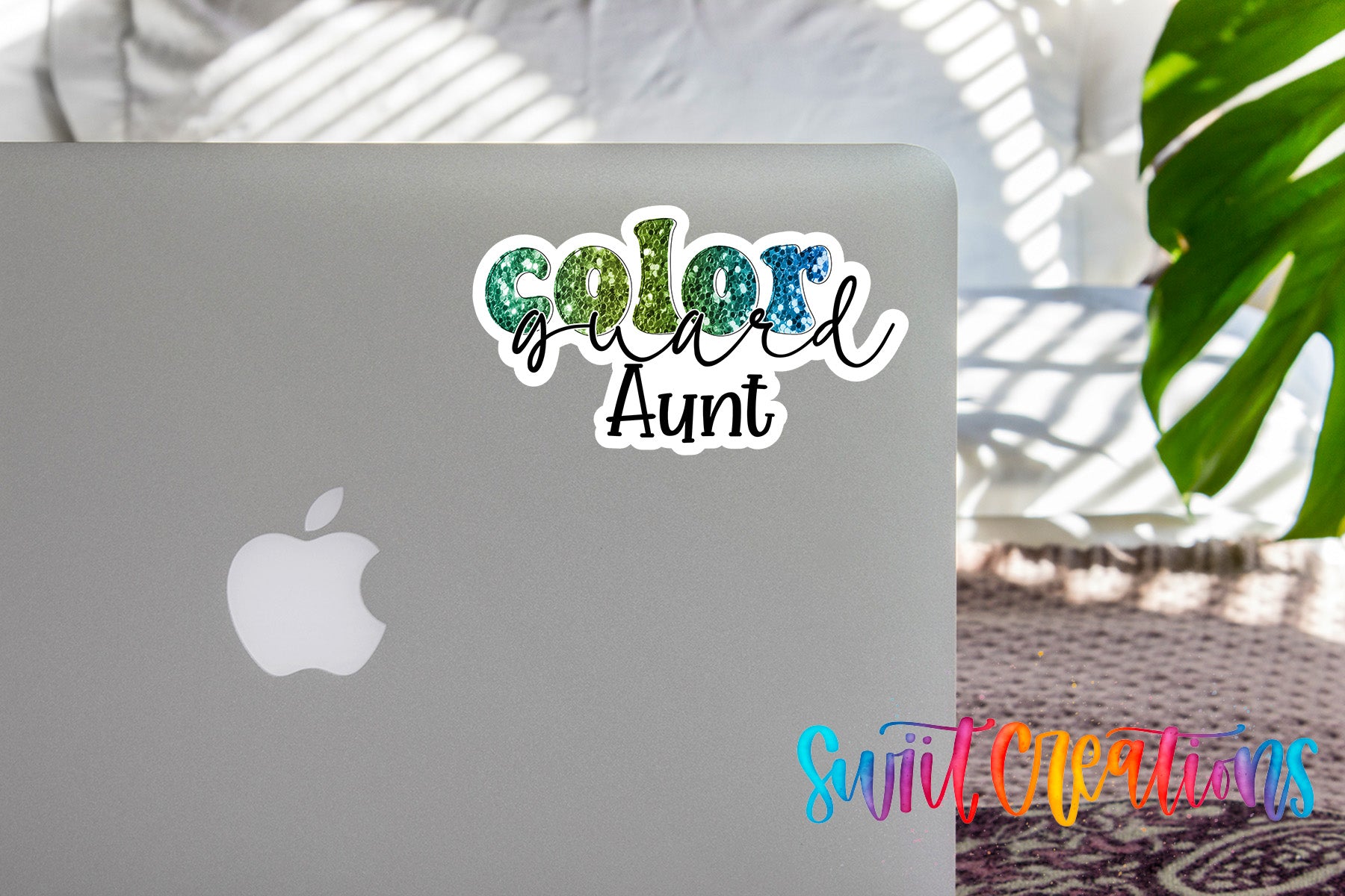 a sticker with the text "Color Guard Aunt" on the back of a silver laptop, with a plant and a patterned rug visible in the background.