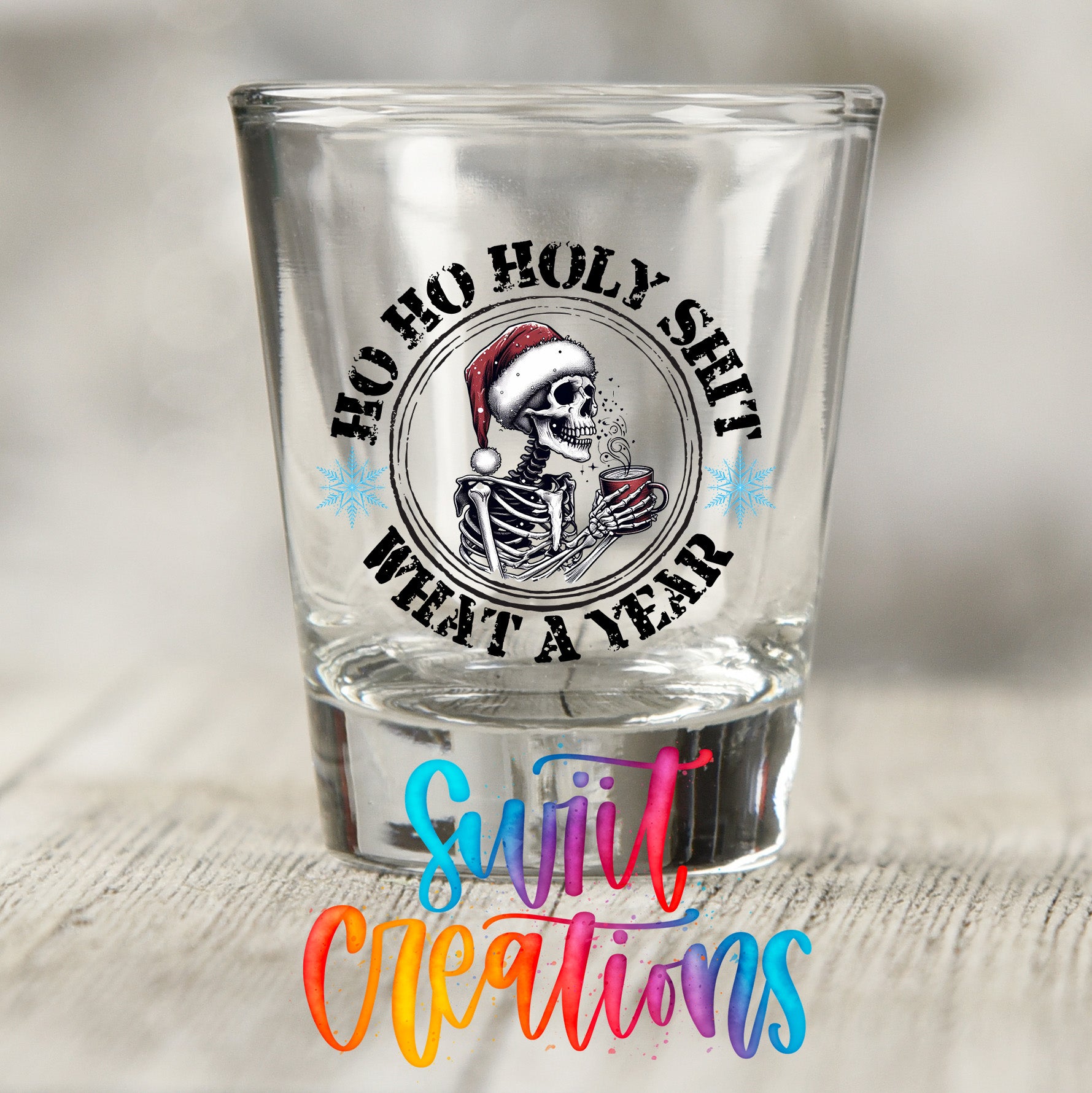 A shot glass with a skull wearing a Santa hat and holding a mug, surrounded by text that reads "Hoo ho holy shit what a year" and "Suit Creations".