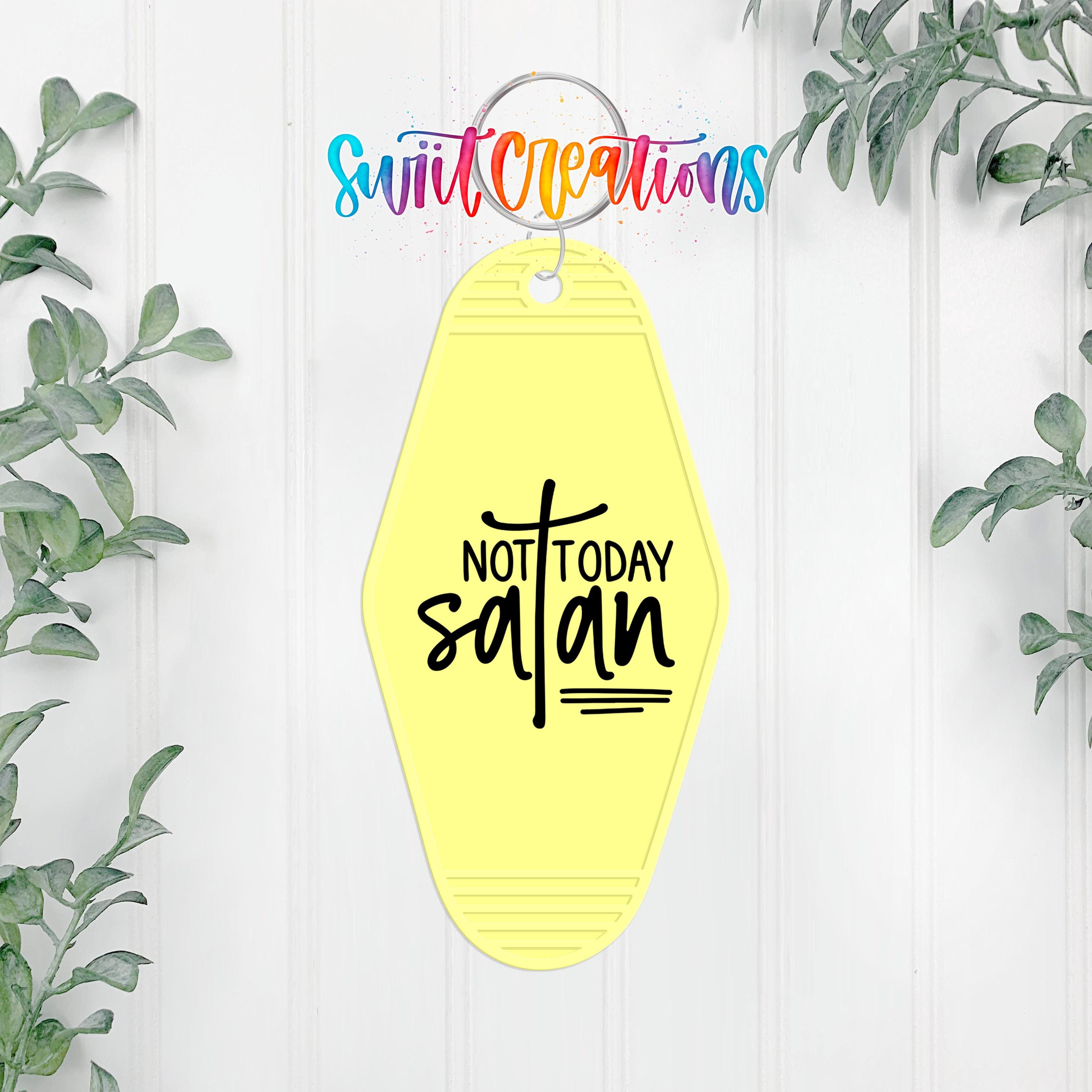 A yellow keychain with the text "NOT TODAY SATAN" printed on it, hanging on a white wall with green plants on either side.